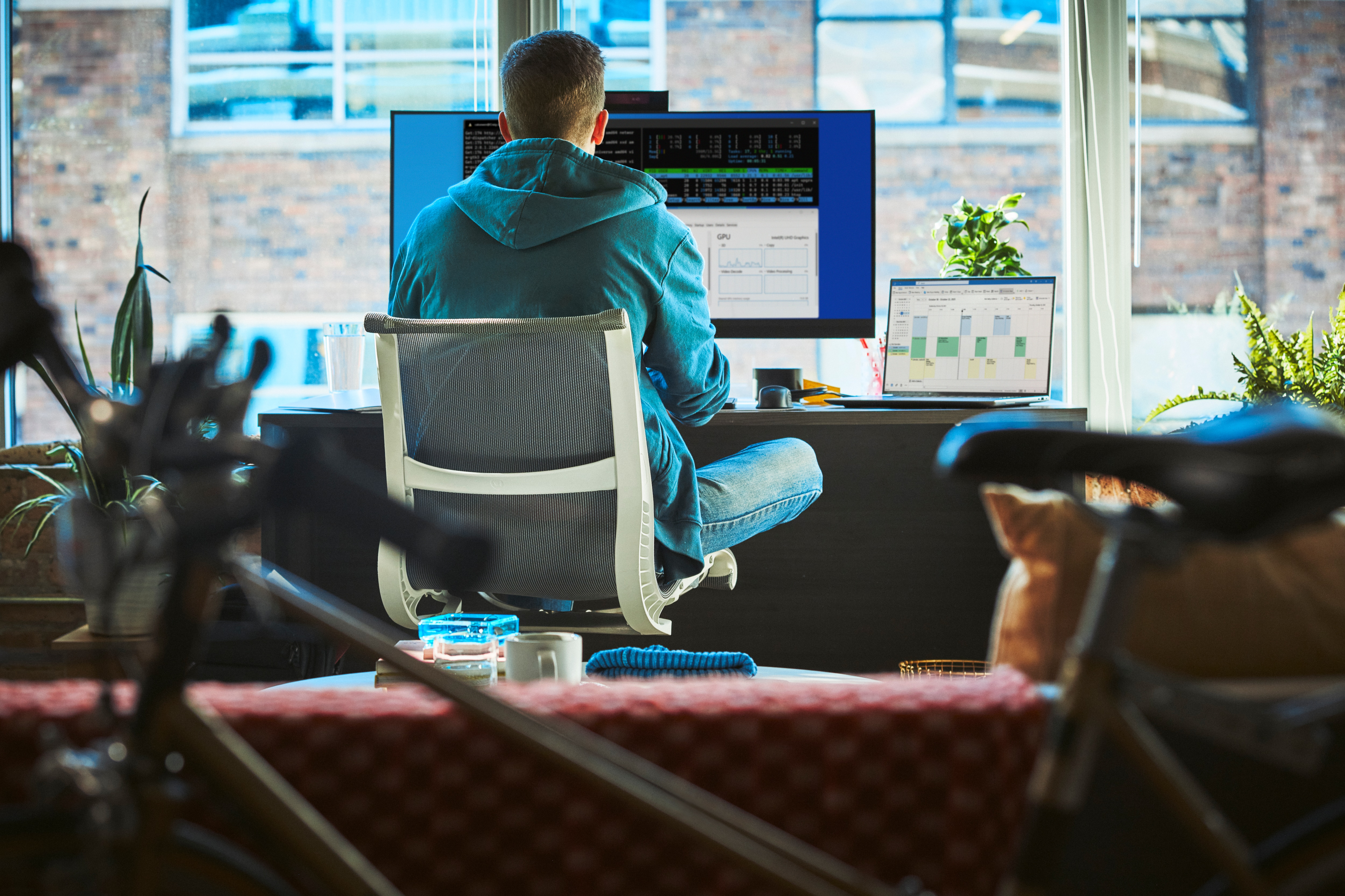Mężczyzna siedzi przy biurku, używając monitora i laptopa. Zawiera klawiaturę, rośliny i widok z okna.