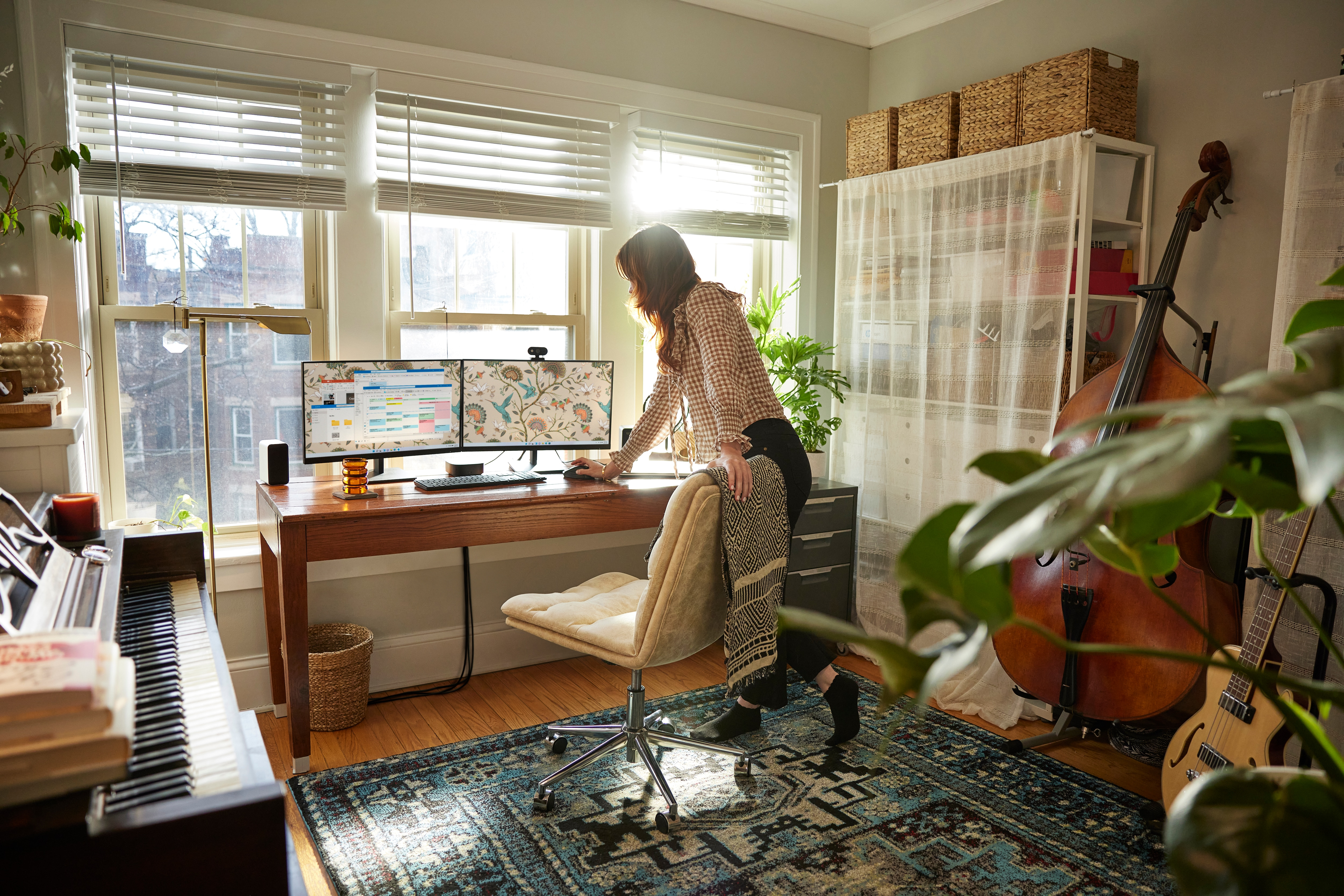 Kobieta pracuje przy biurku z dwoma monitorami, pianinem i wiolonczelą w pokoju.