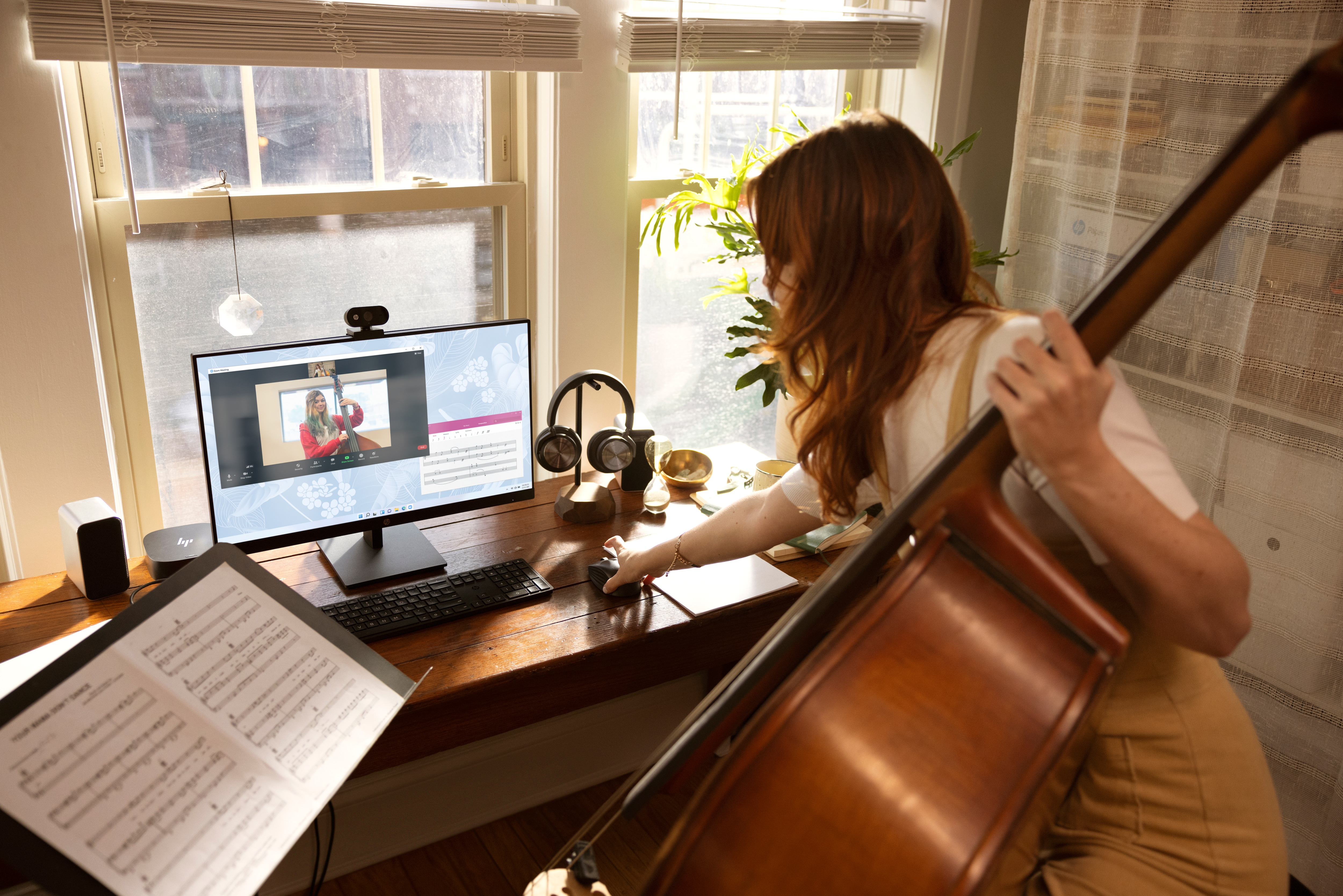 Mujer en escritorio con monitor, violonchelo y partituras durante videollamada.
