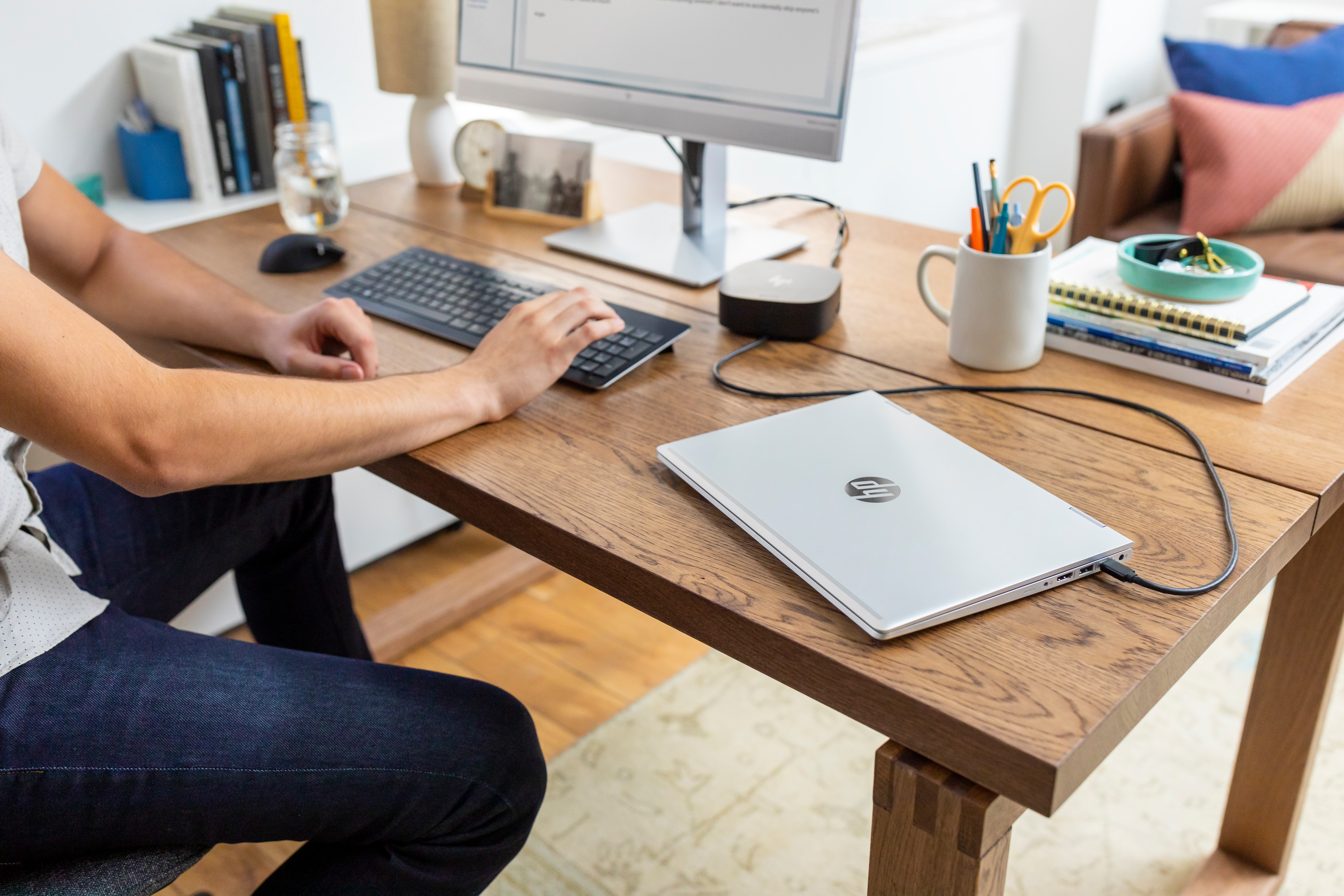 Persona escribiendo en un teclado en un escritorio de madera con un ordenador y un portátil. Otros objetos también están en el escritorio.