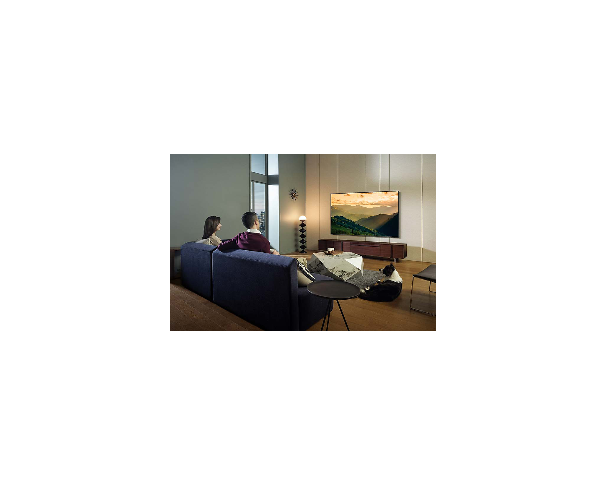 Para siedzi na niebieskiej kanapie, naprzeciwko telewizora z krajobrazem.