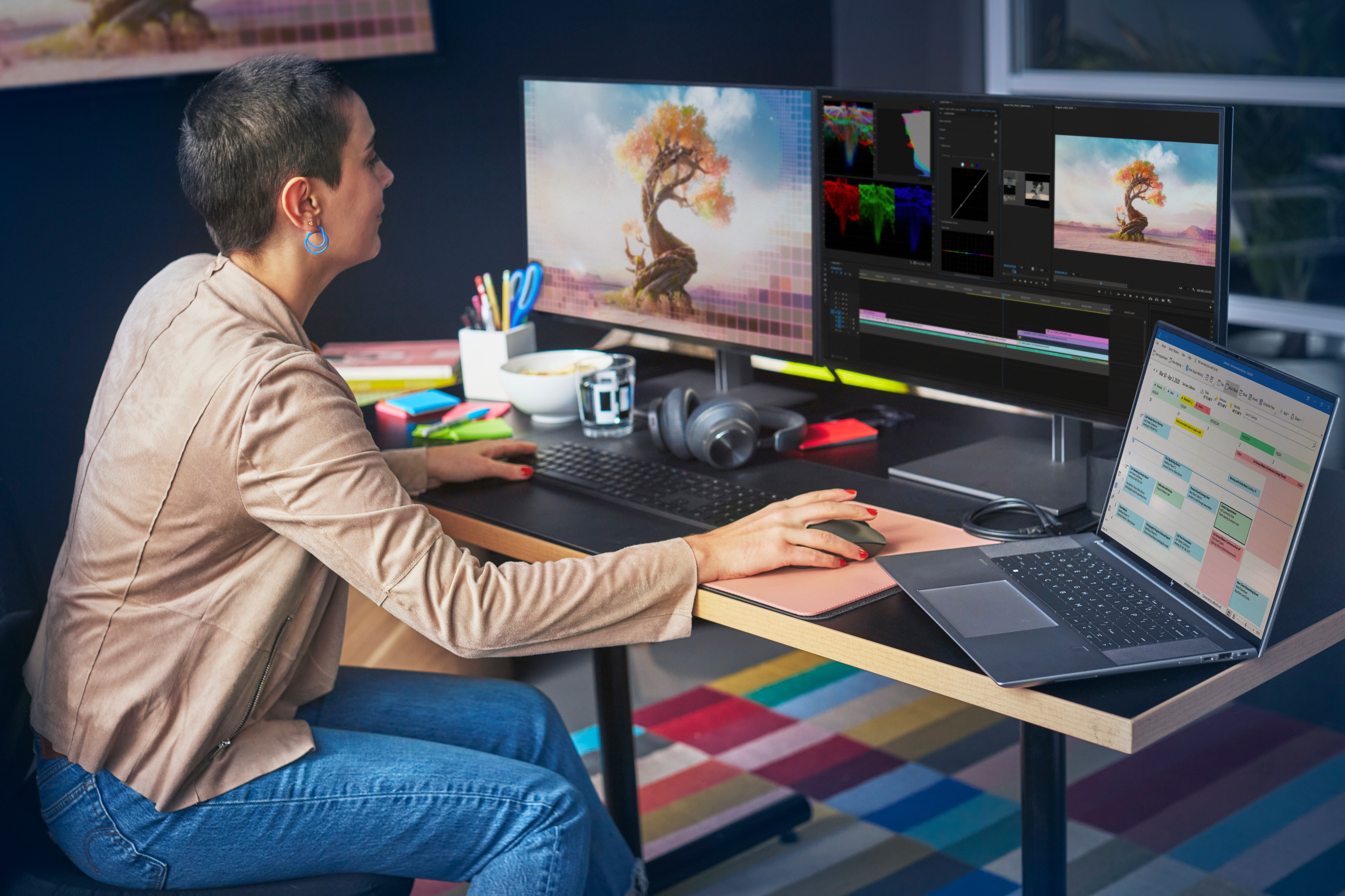 Kobieta siedzi przy biurku z konfiguracją komputera. Są dwa monitory i laptop.