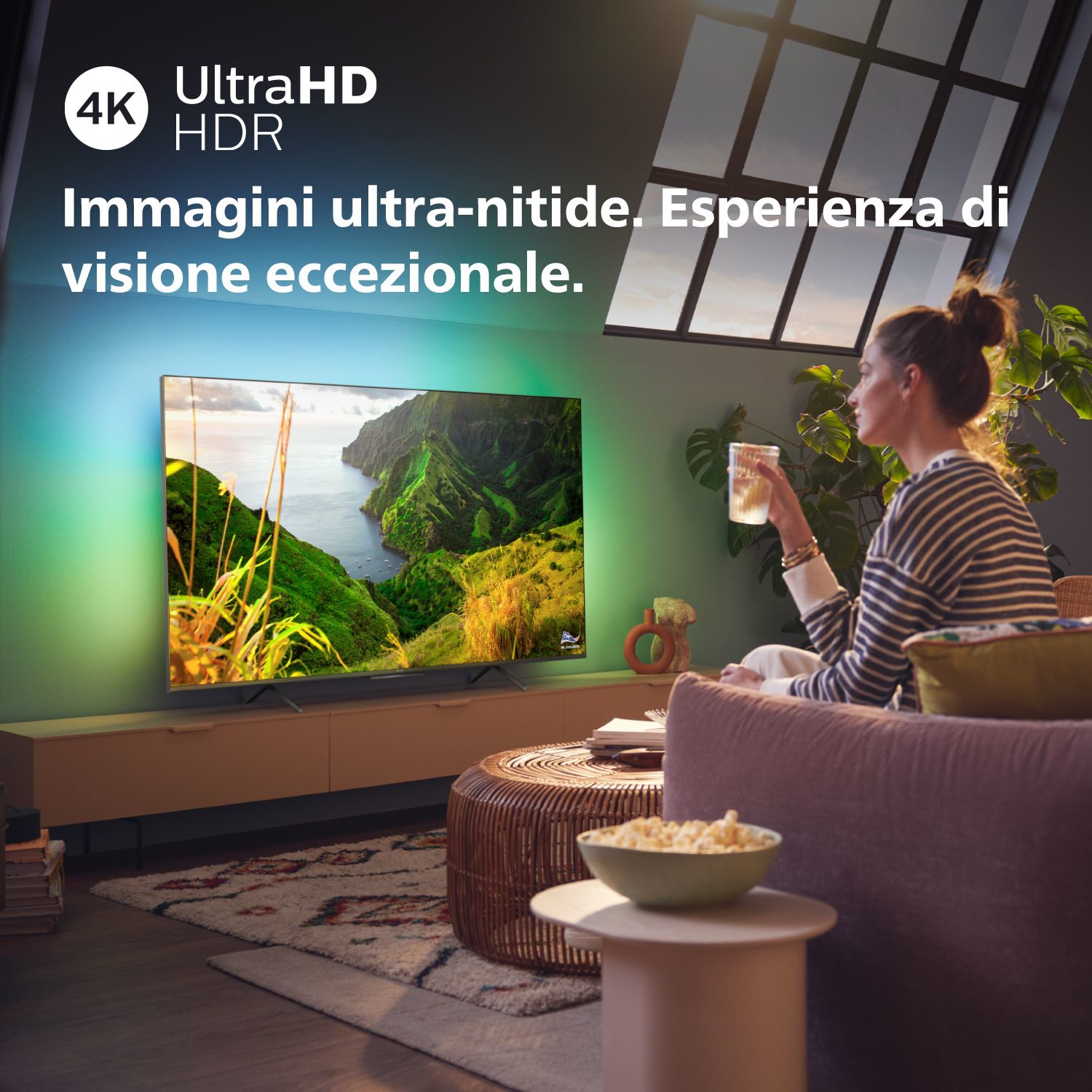 Una mujer está sentada en un sofá viendo la televisión que muestra un paisaje. Hay palomitas de maíz.