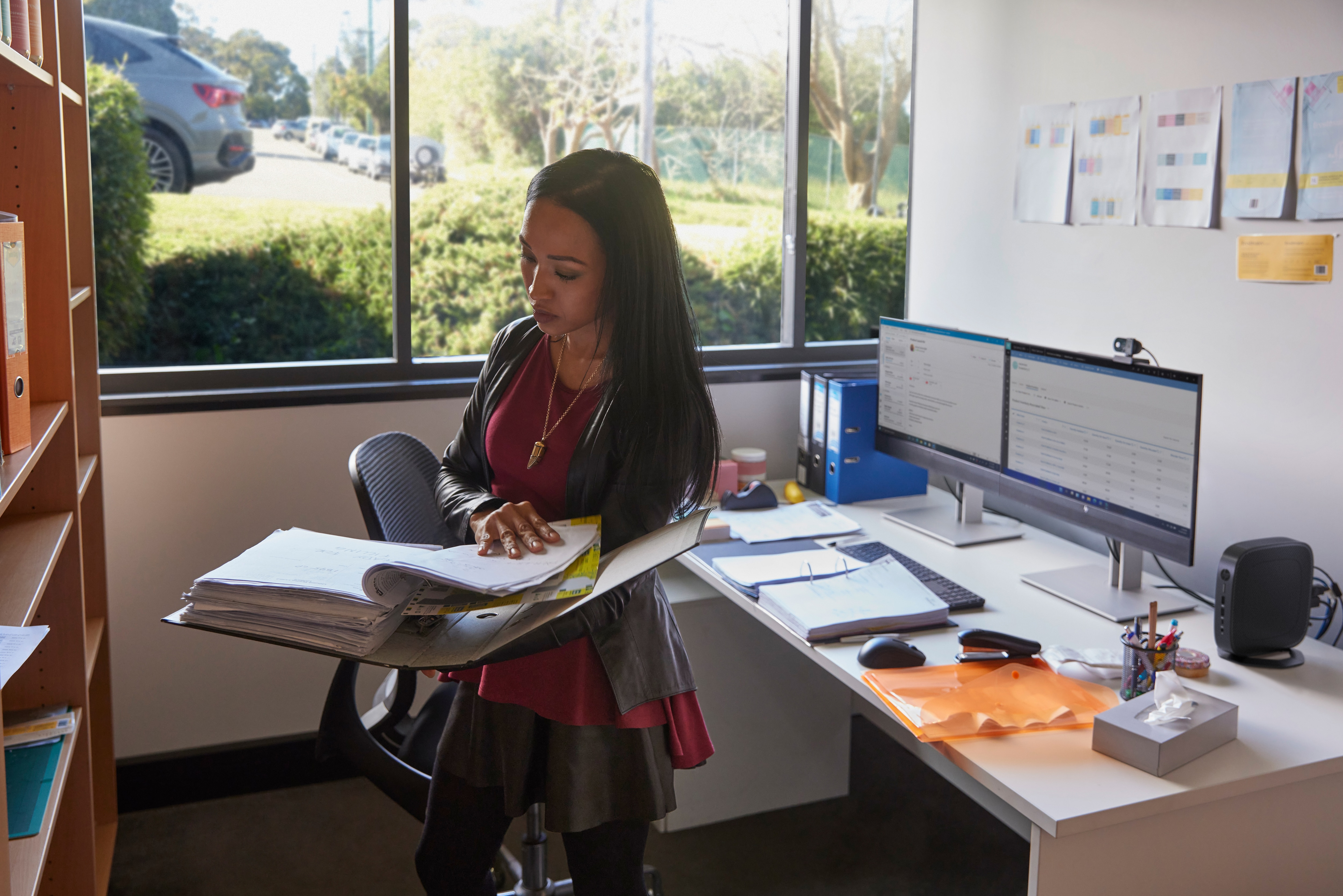 Kobieta w biurze trzymająca dokumenty, z monitorami, biurkiem i regałem z książkami w tle.