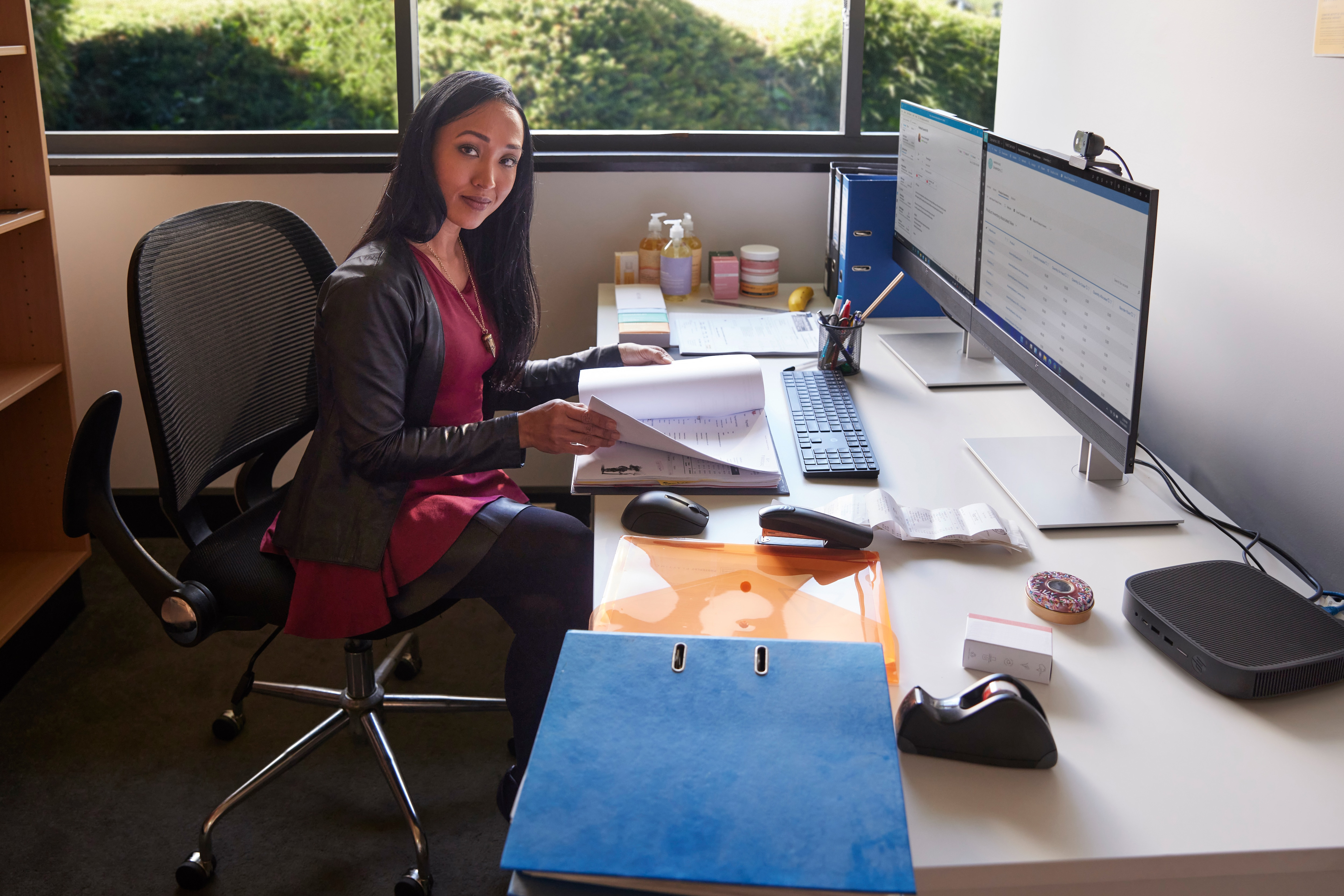 Kobieta siedzi przy biurku z dwoma monitorami, trzymając dokumenty. Jest klawiatura, mysz i inne przedmioty.