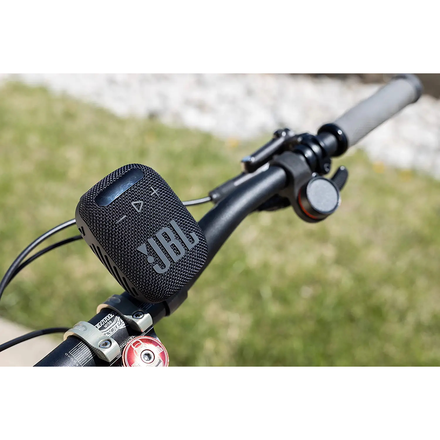 Schwarzer JBL-Lautsprecher an Fahrradlenkern montiert. Der Hintergrund zeigt grünes Gras und Sonnenlicht.