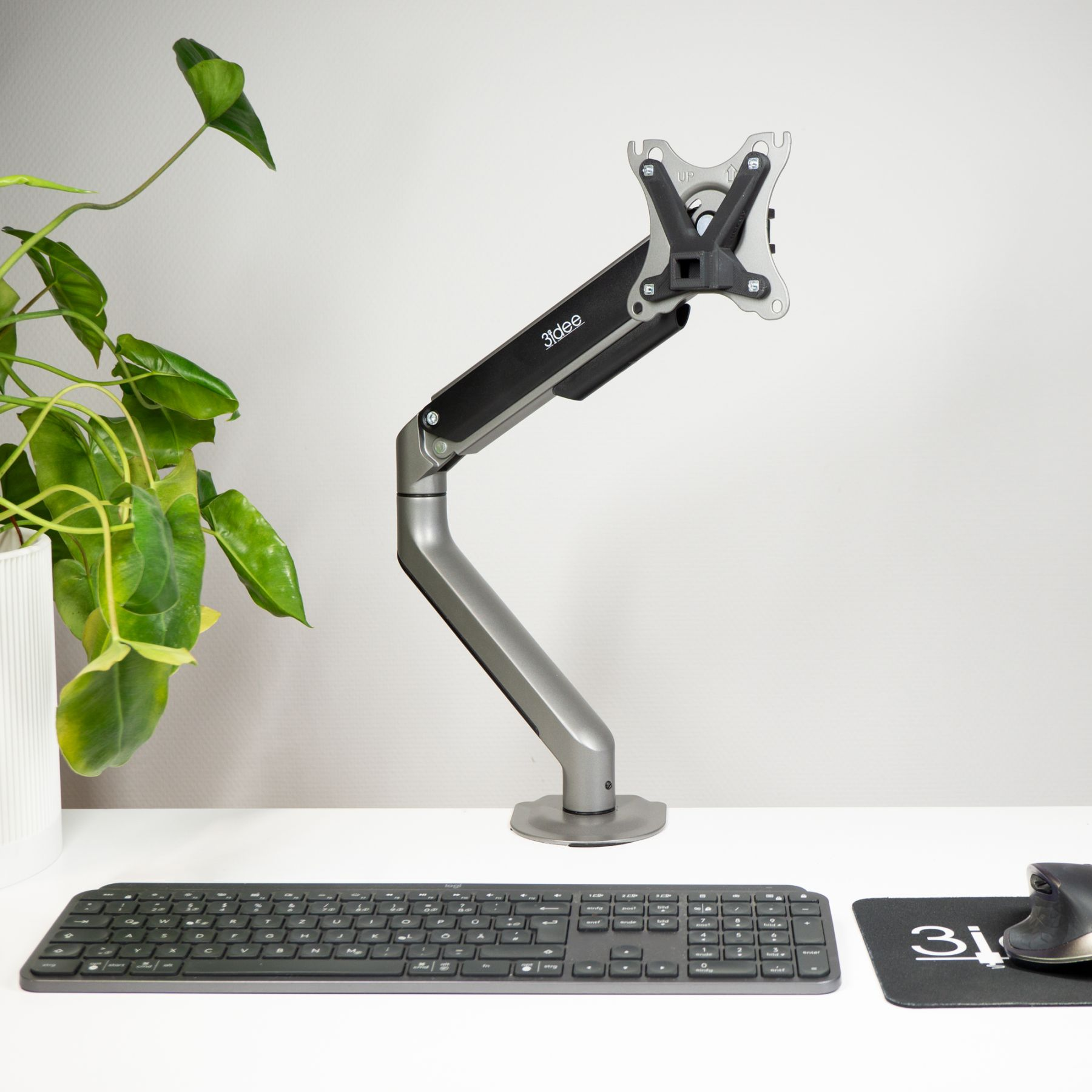 Monitorarm. Der Arm ist silber und schwarz. Eine schwarze Tastatur und Maus darunter. Eine grüne Pflanze links.