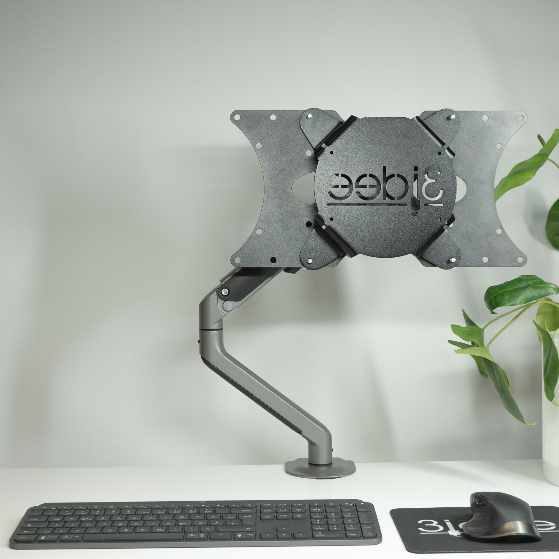 Ein Monitor und ein Monitorarm auf einem Schreibtisch, mit Tastatur, Maus und weißer Wand im Hintergrund.