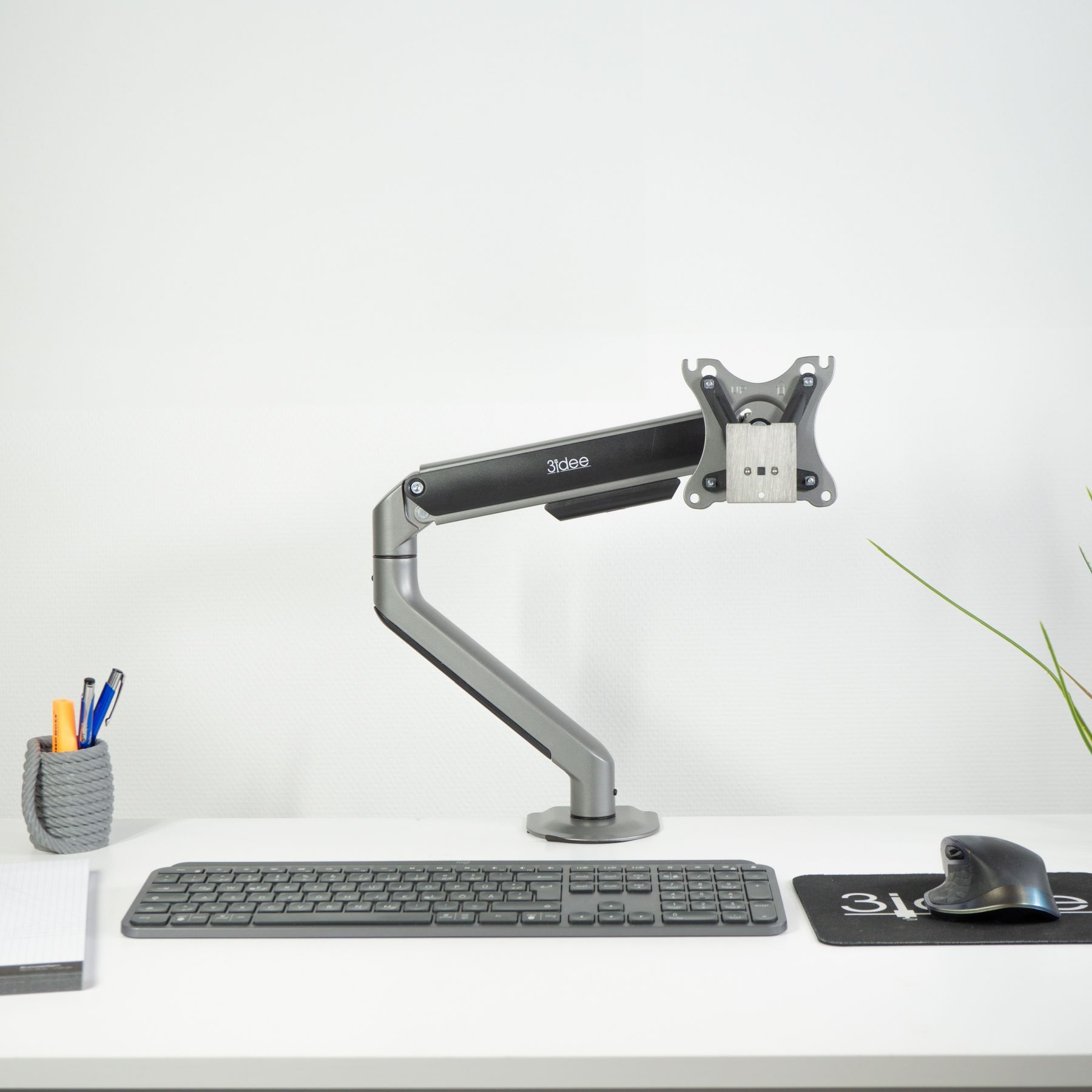 Monitorarm auf weißem Schreibtisch mit Tastatur, Maus und Büromaterial. Weiße Wand dahinter.