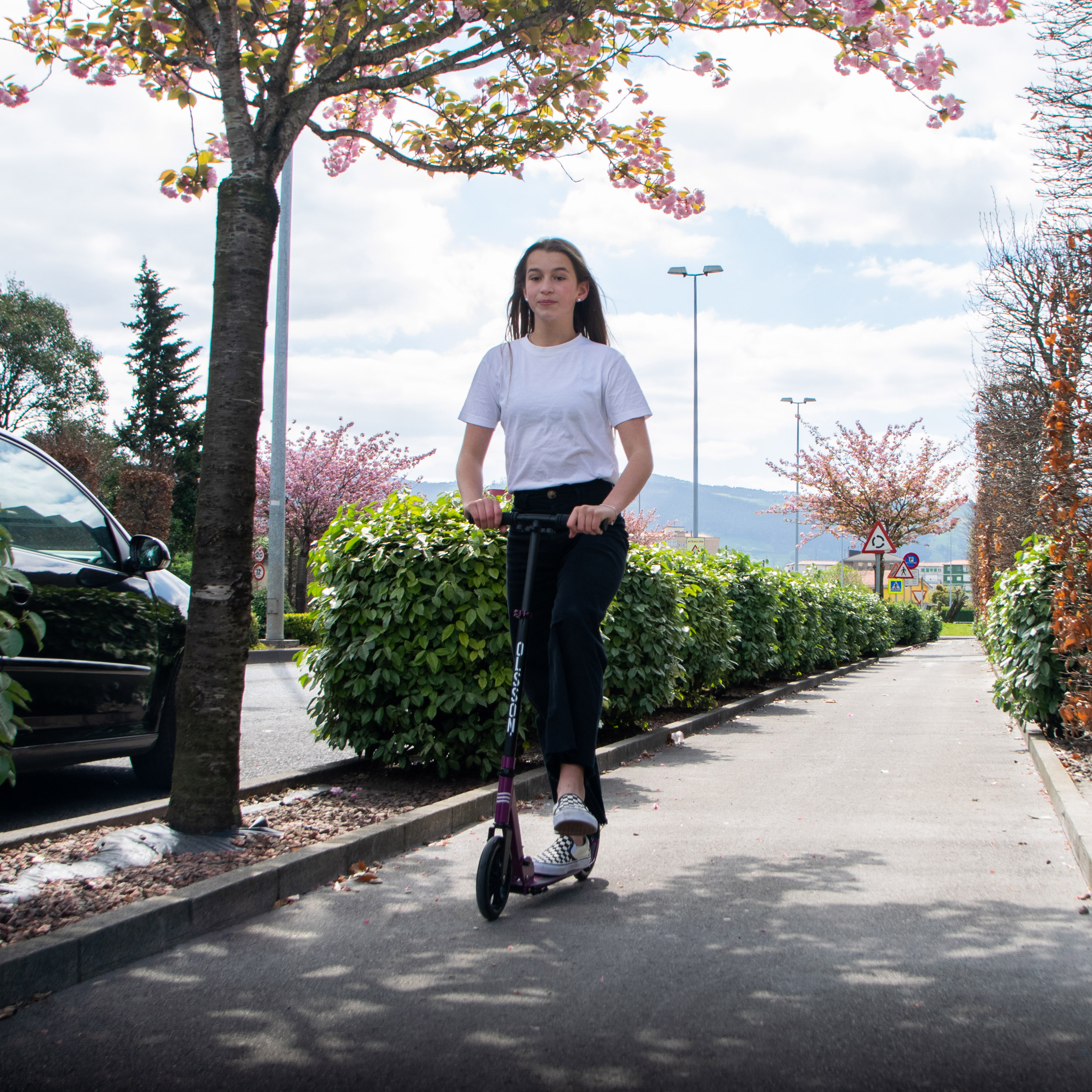 Joven montada en un patinete morado en una acera, junto a un seto y una carretera.