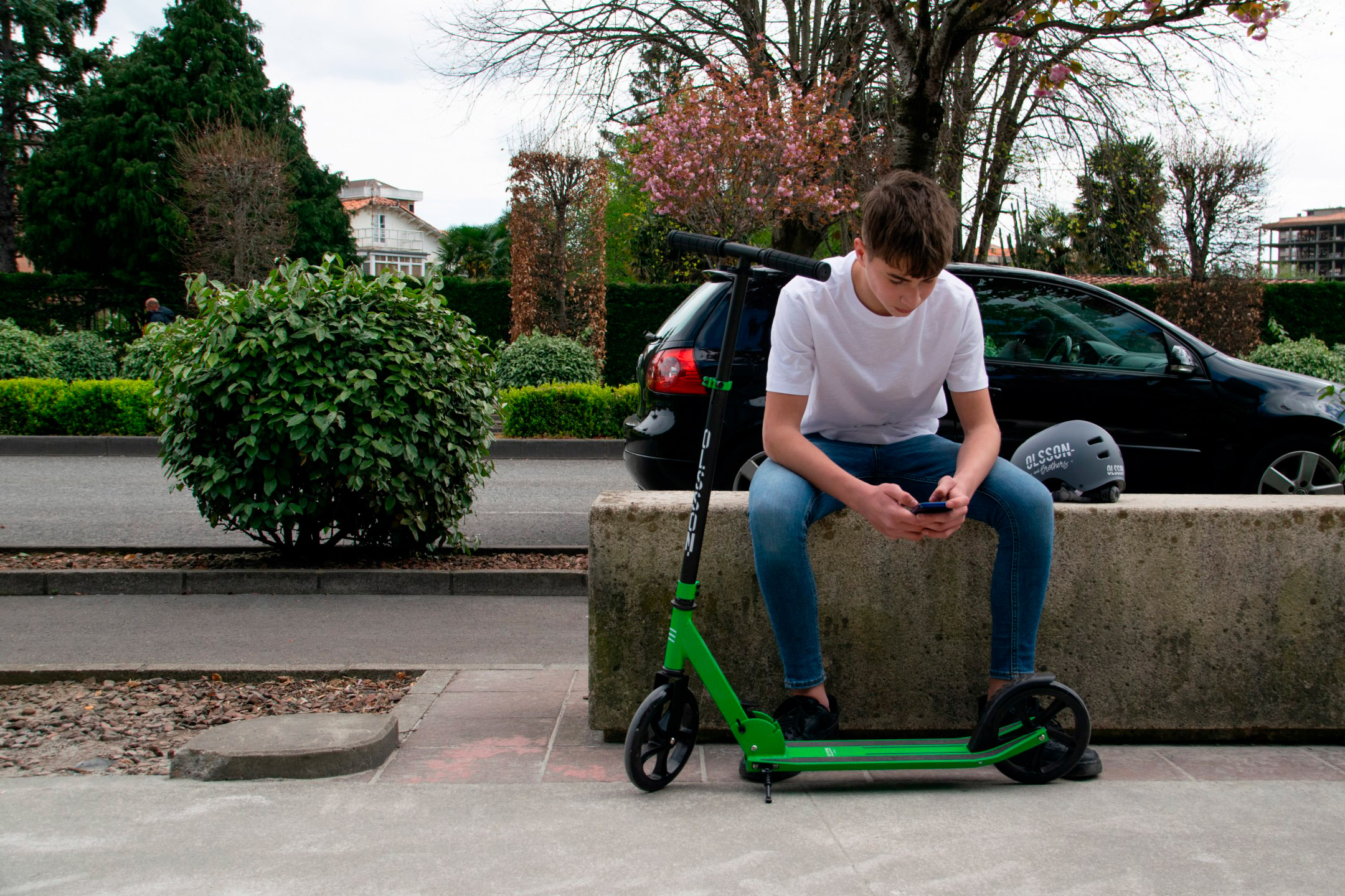 Un joven sentado en un muro usando su teléfono, con un patinete verde al lado.