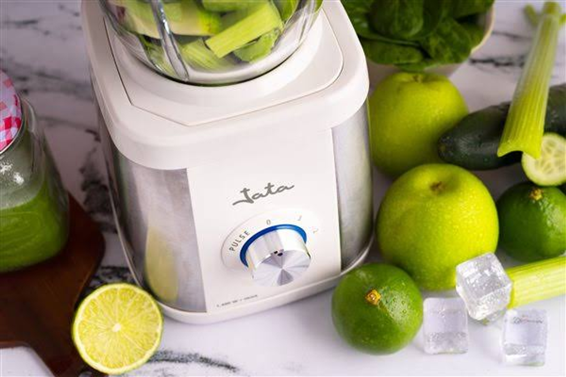 Blender z selerem w środku, jabłka, limonki, kostki lodu i połówka cytryny. Białe tło.