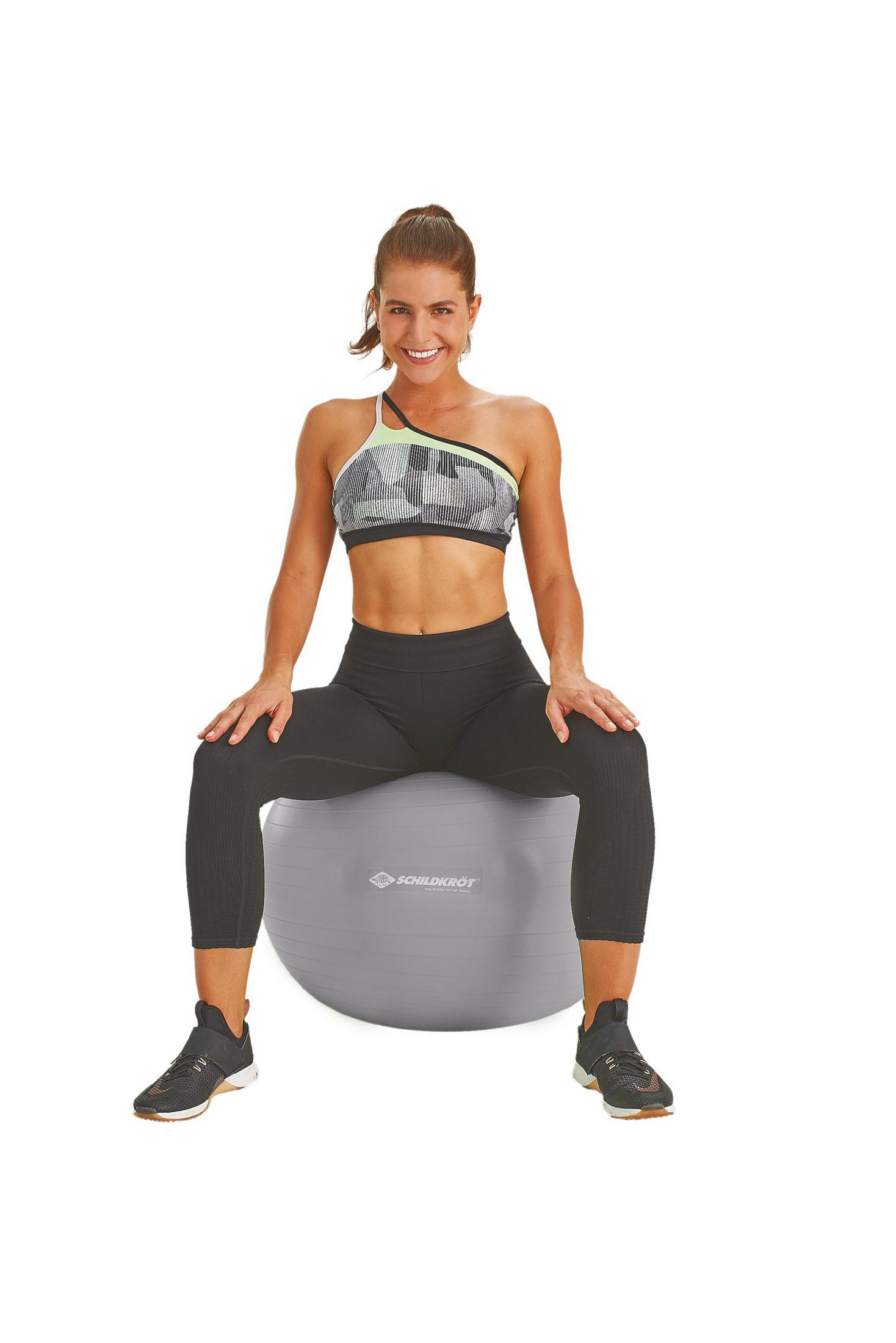 Frau auf grauem Gymnastikball. Sport-BH und Leggings. Weißer Hintergrund.