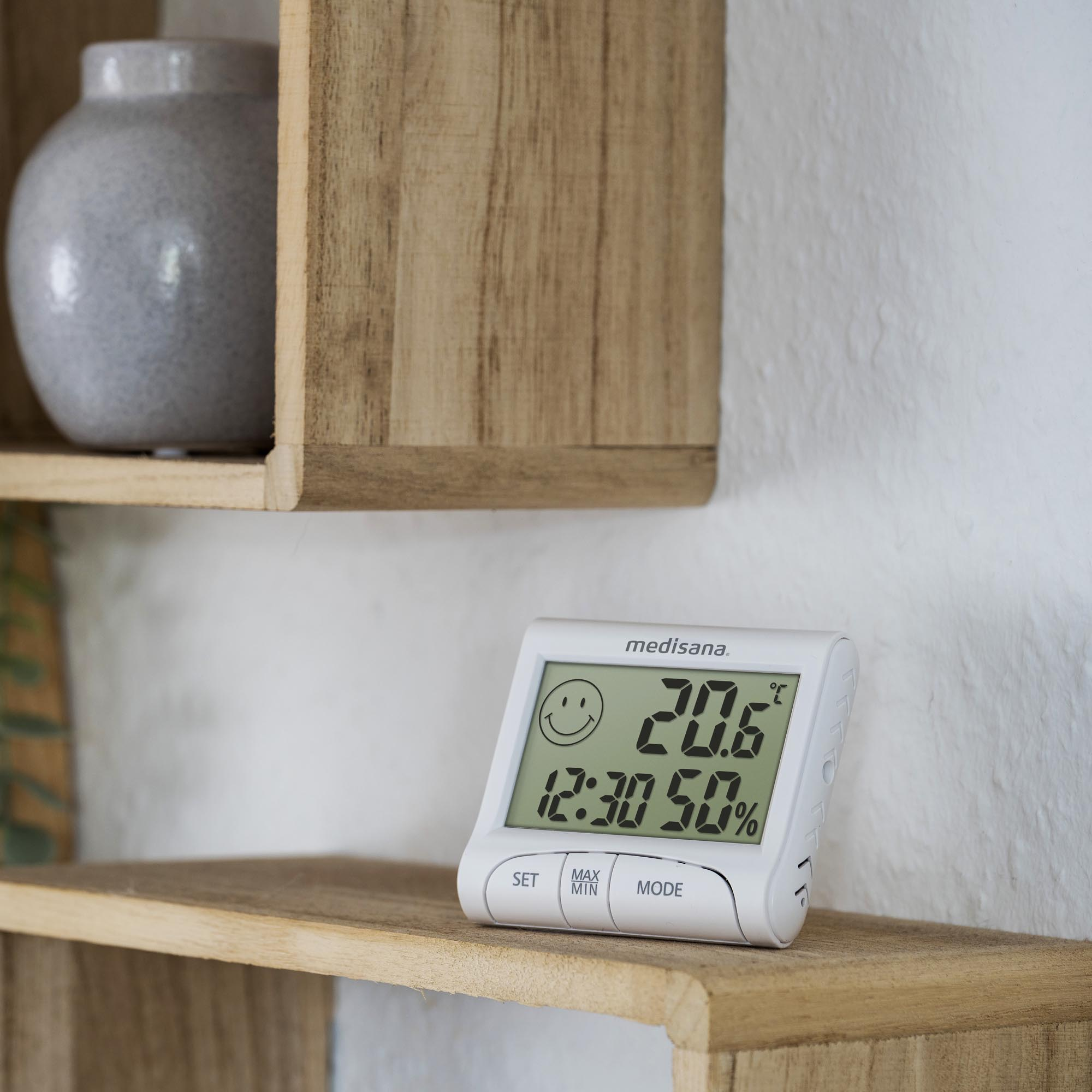 Weißes digitales Thermometer auf einem Holzregal. Es zeigt Temperatur und Luftfeuchtigkeit mit einem Smiley an.