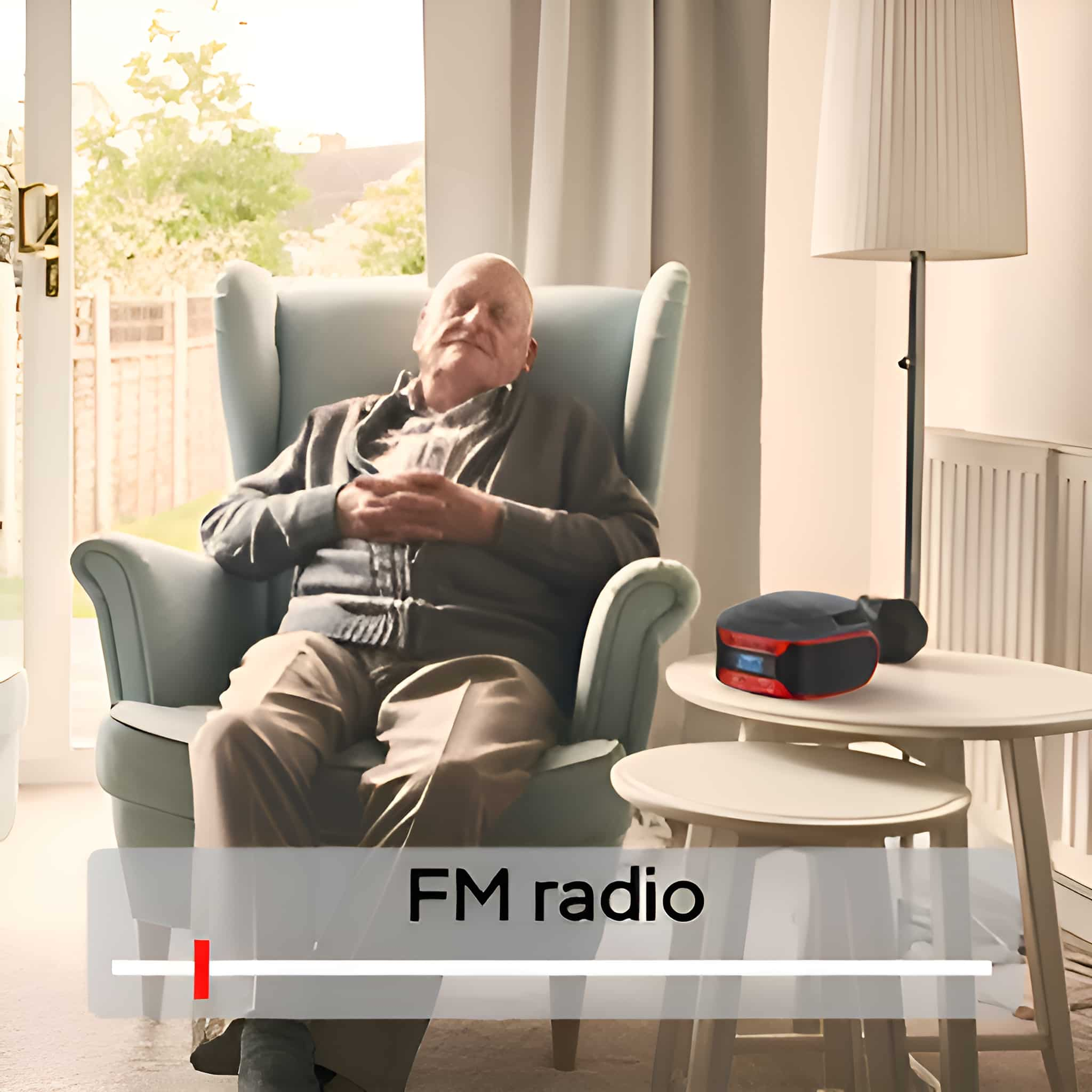 Ein alter Mann sitzt in einem Stuhl mit einem Radio auf einem Tisch. Der Hintergrund ist ein Raum mit einem Fenster.
