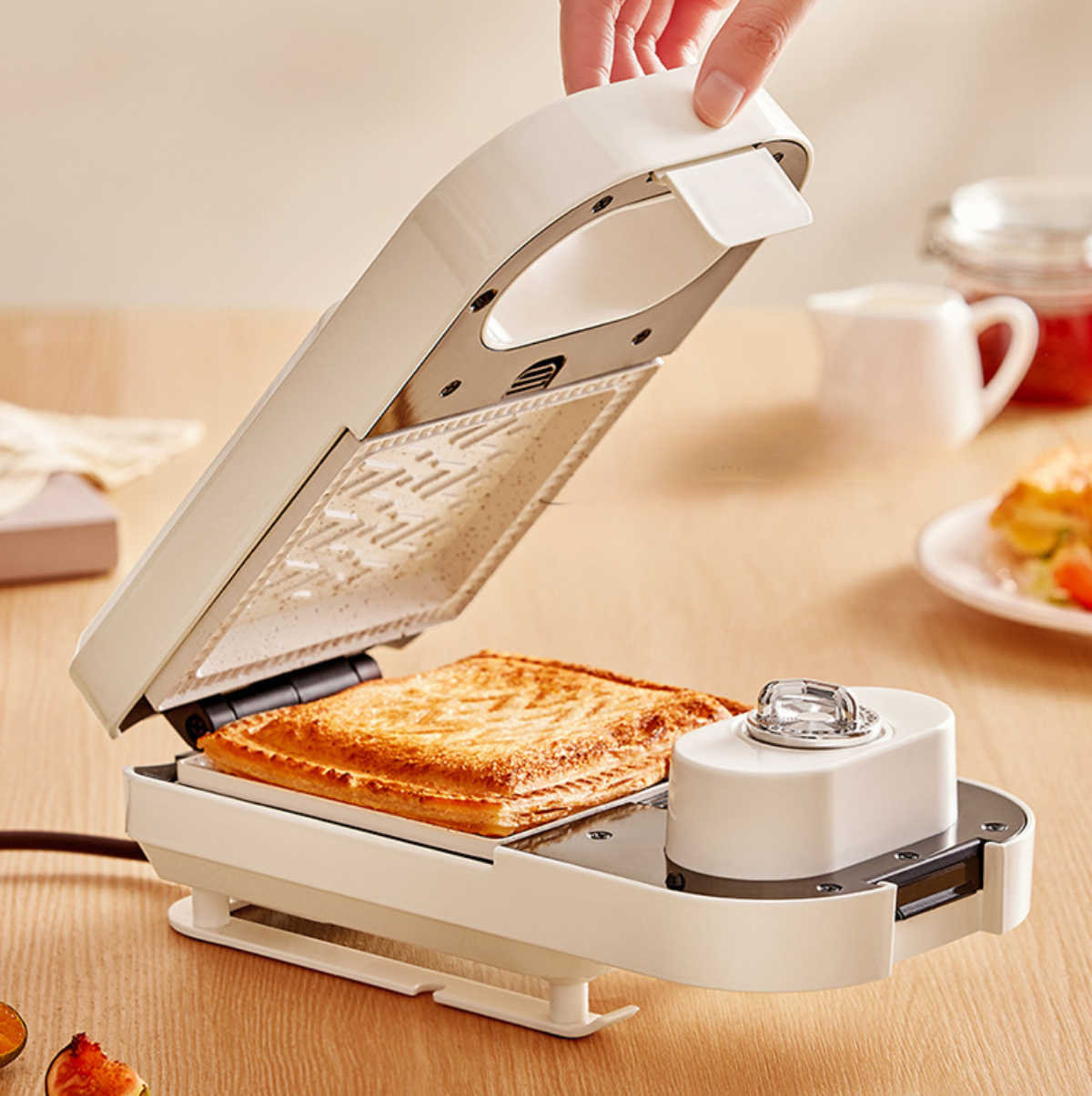 Ein weißer Sandwichmaker mit einem getoasteten Sandwich darin, das von einer Hand geöffnet wird. Andere Lebensmittel sind dabei.