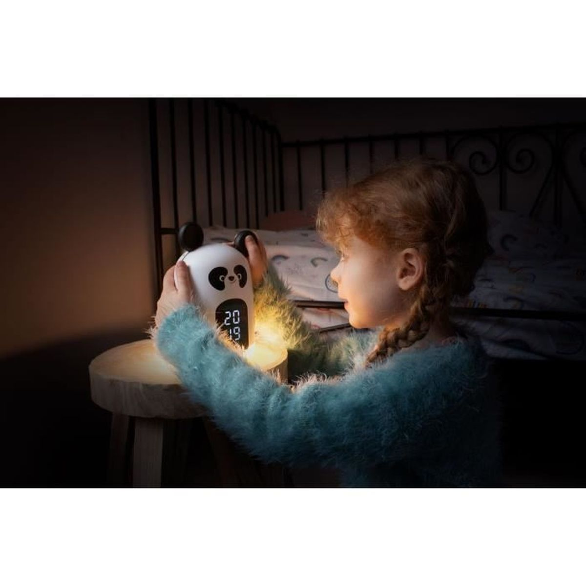Dziewczynka w niebieskim swetrze patrzy na budzik w kształcie pandy na drewnianym stołku w ciemnym pokoju.
