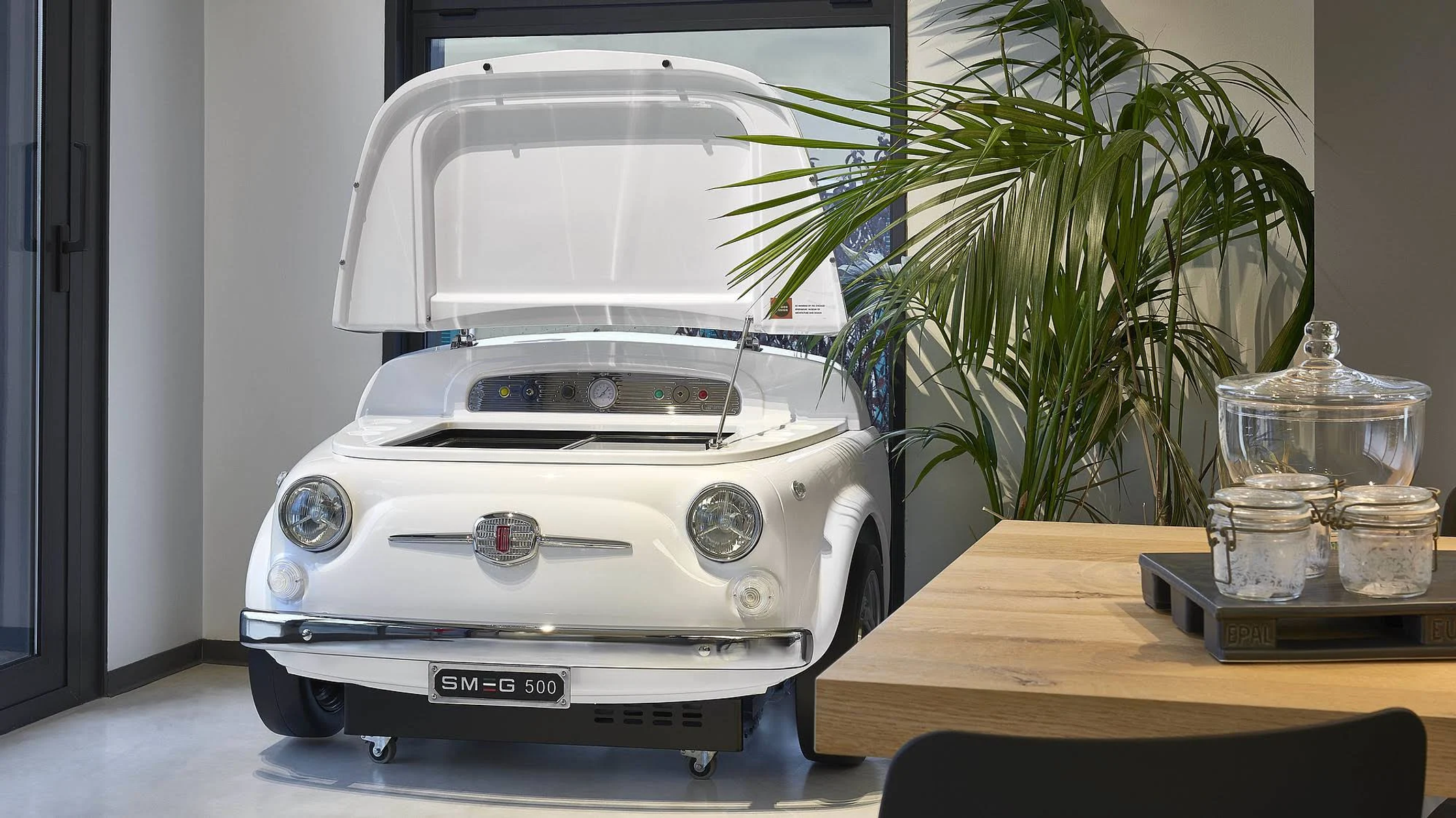 Coche blanco con capó abierto dentro de un edificio. Hay una mesa y plantas.