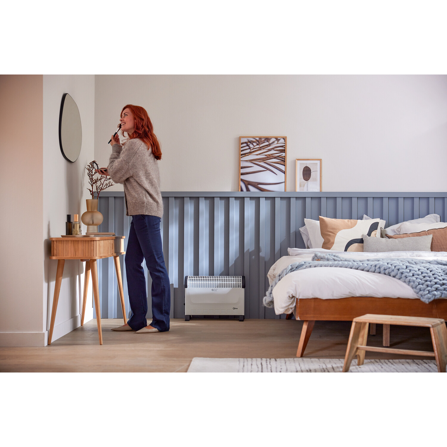 Kobieta nakładająca makijaż, sypialnia. Toaletka, lustro, łóżko z poduszkami, grzejnik, drewniana podłoga.