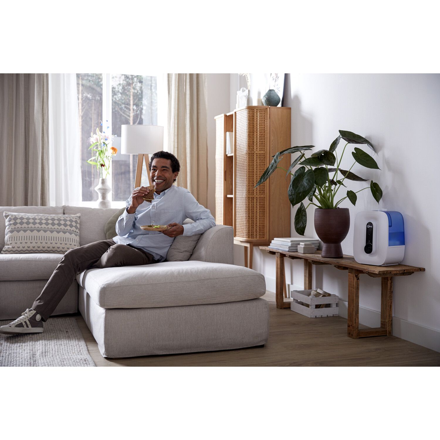 Mężczyzna siedzi na sofie i je. W pobliżu znajduje się roślina i nawilżacz powietrza na stole.