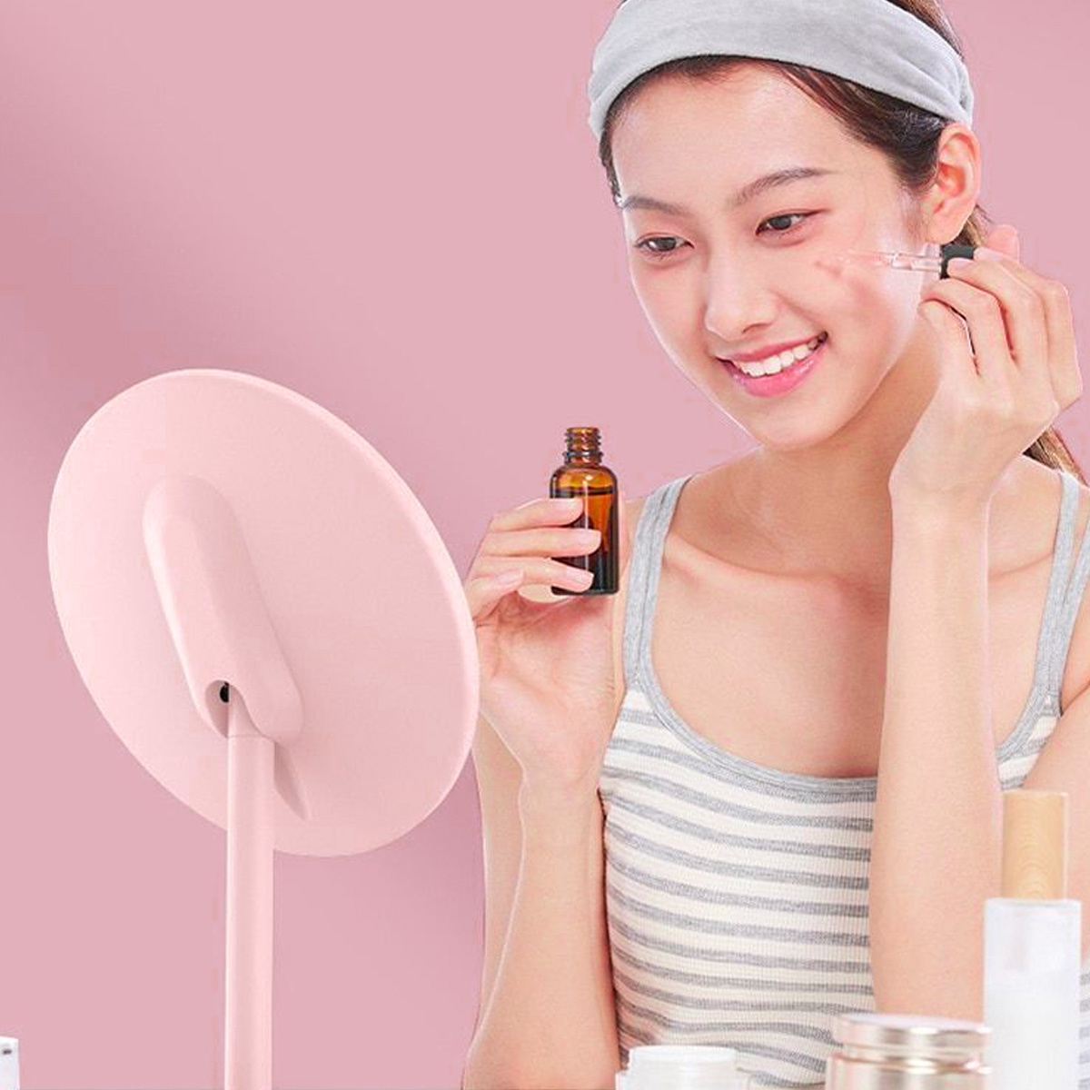 Frau trägt Serum von einer Pipette auf, während sie in einen rosa Spiegel schaut. Ein rosa Hintergrund ist vorhanden.