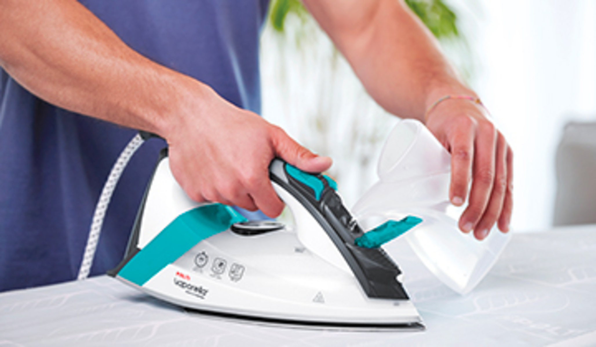 Osoba prasuje białym i turkusowym żelazkiem. Dolewa wodę.