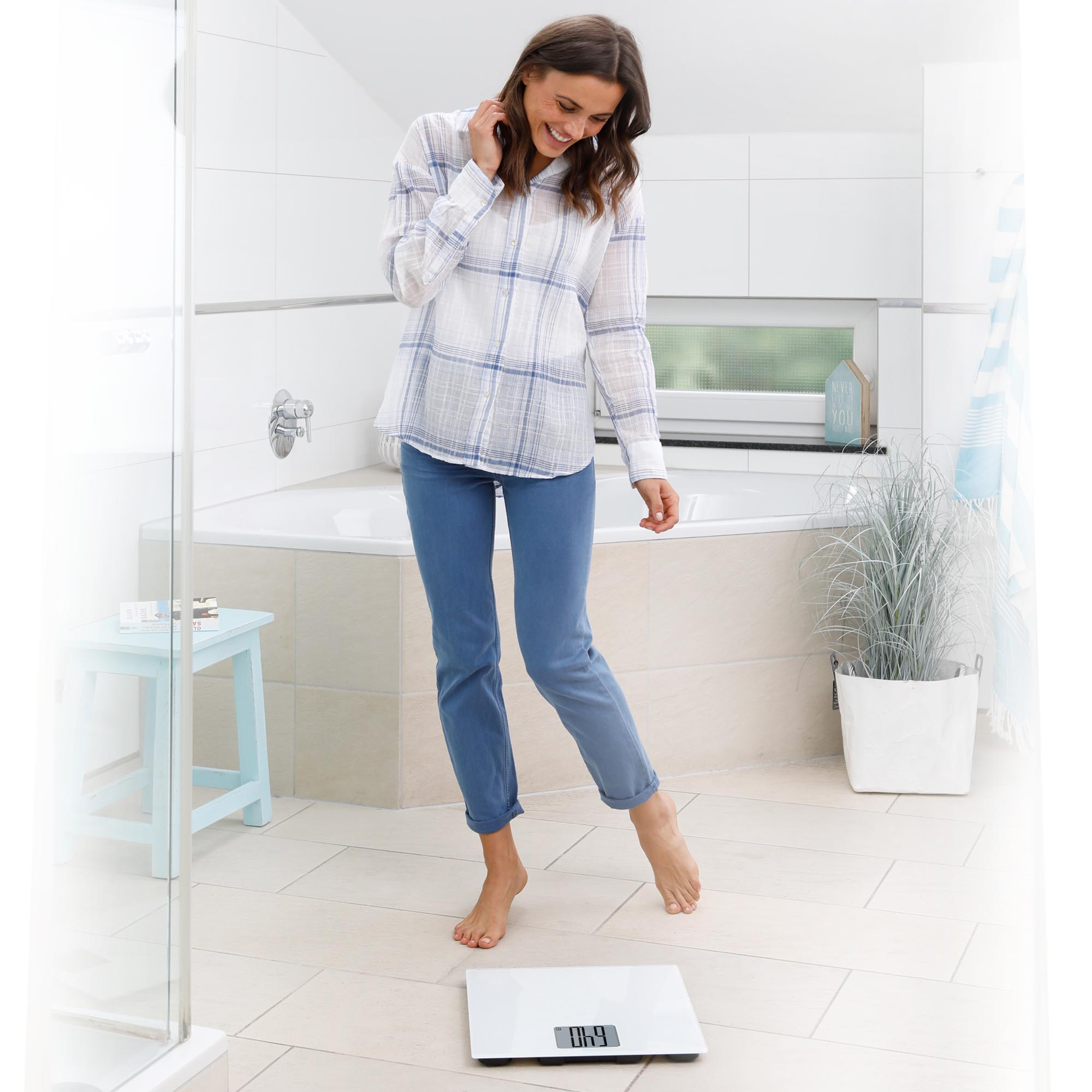 Kobieta stoi na wadze w łazience. Uśmiecha się, w dżinsach i koszuli. Biała wanna i stołek w tle.
