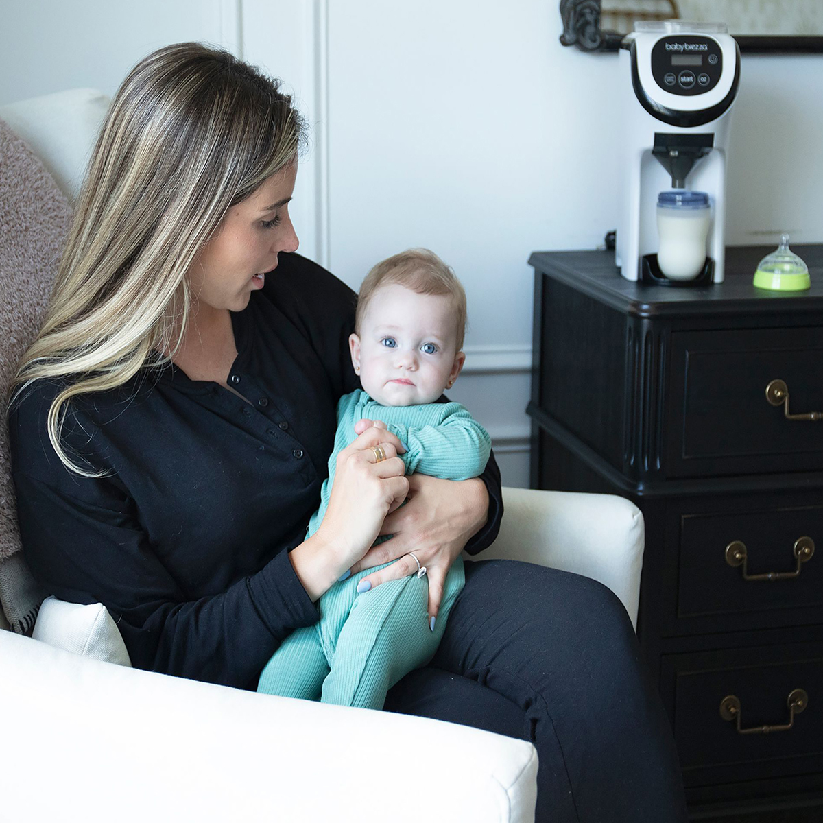 Frau hält ein Baby, sitzt in einem Stuhl, in der Nähe einer Babynahrungsmaschine.