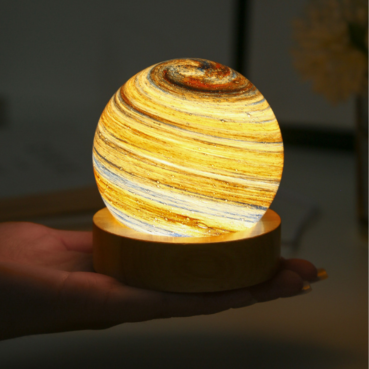 Eine beleuchtete dekorative Lampe in Planetenform auf einem Holzsockel, die in einer Hand gehalten wird.