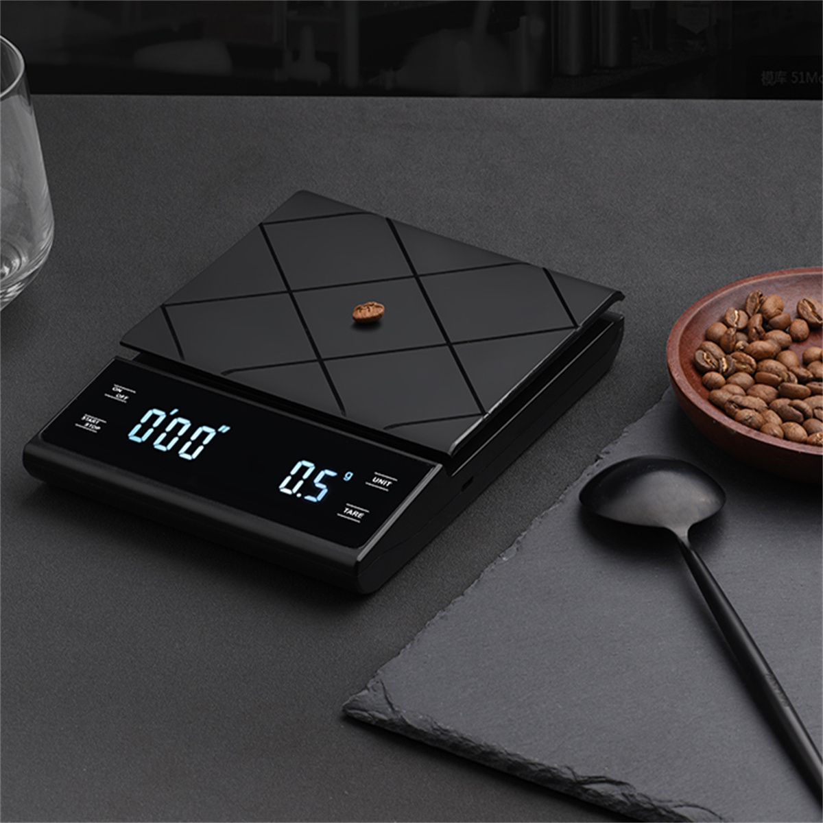 Eine schwarze Waage mit einer Kaffeebohne darauf. Kaffeezubehör und die Waage stehen auf einem Tisch.