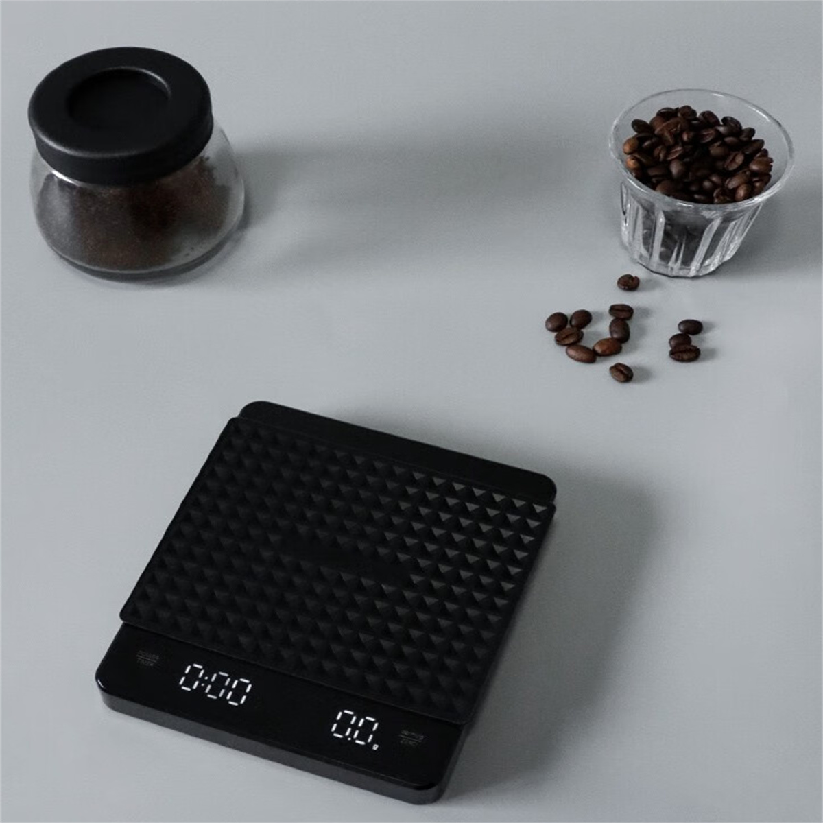Eine schwarze Digitalwaage mit Kaffeebohnen und gemahlenem Kaffee in Gläsern auf grauer Oberfläche.