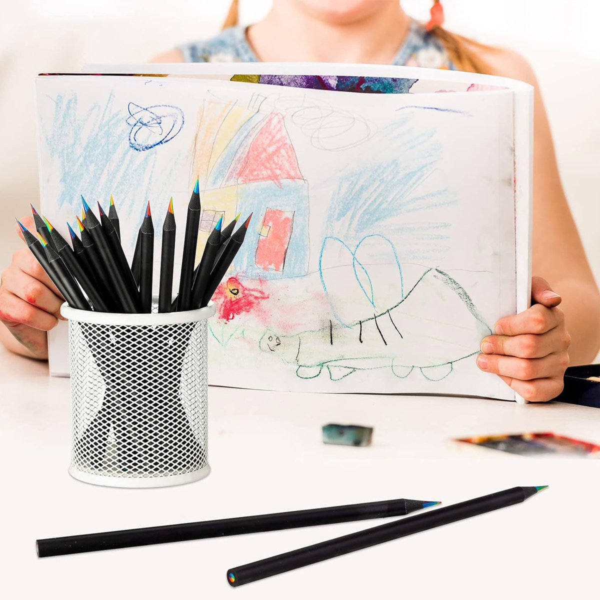 Buntstifte in einem weißen Metallbecher, mit einer Kinderzeichnung im Hintergrund. Zwei Stifte liegen auf dem Tisch.