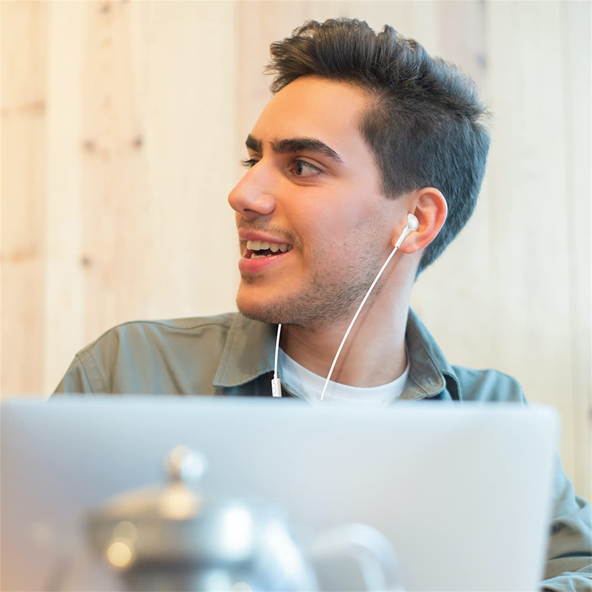 Ein junger Mann mit Ohrhörern lächelt, arbeitet an einem Laptop, Tasse und Teekanne daneben.