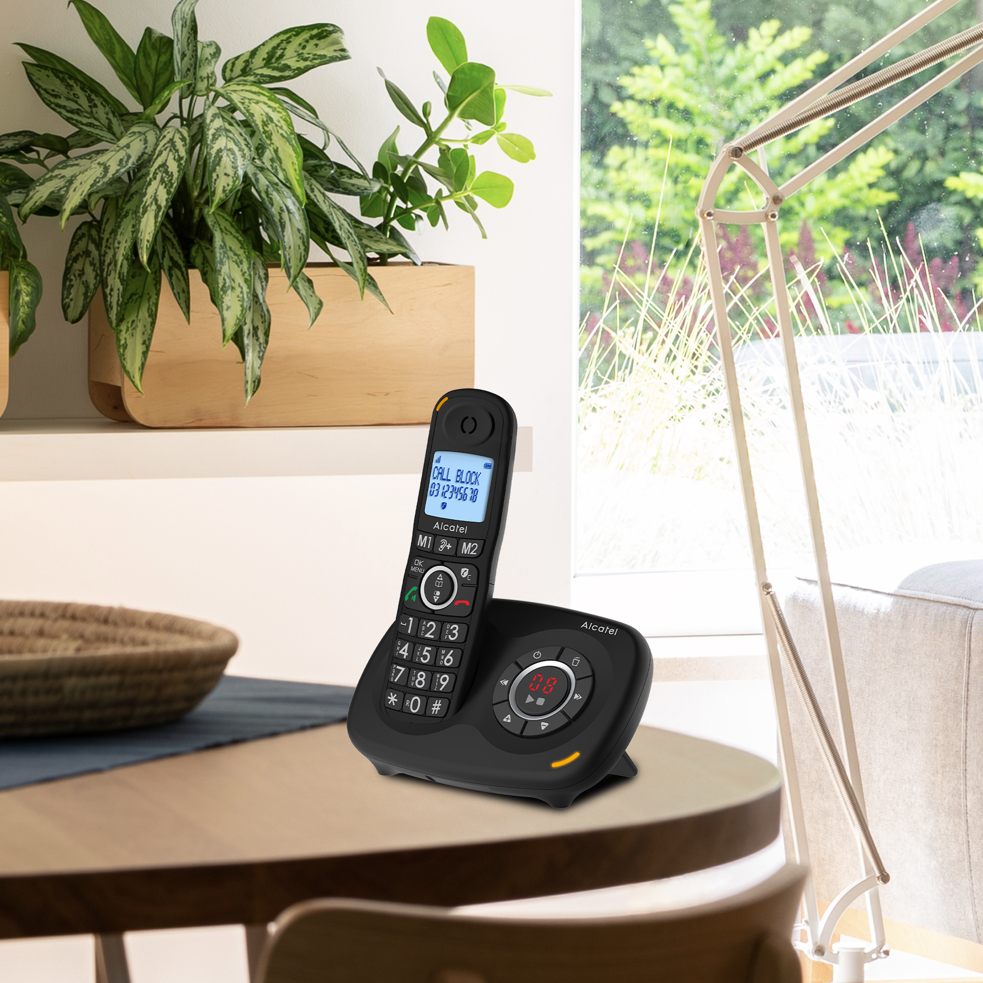 Telefono cordless su un tavolo. Pianta e finestra sullo sfondo. Telefono nero con display acceso. Tavolo beige.