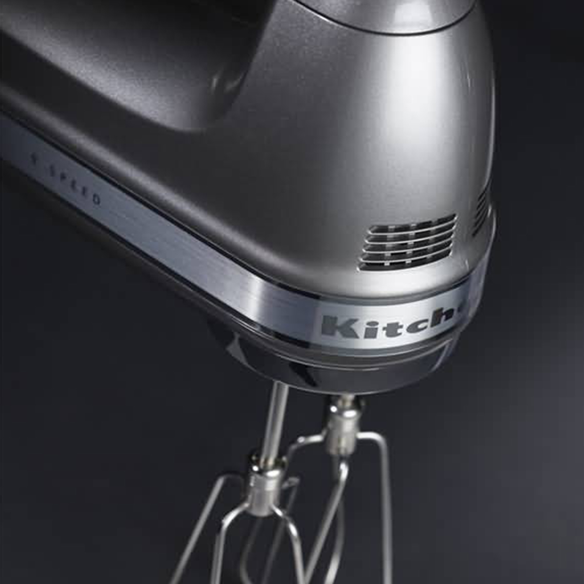 Un frullatore a mano KitchenAid argentato con fruste. Lo sfondo è scuro e sfocato.