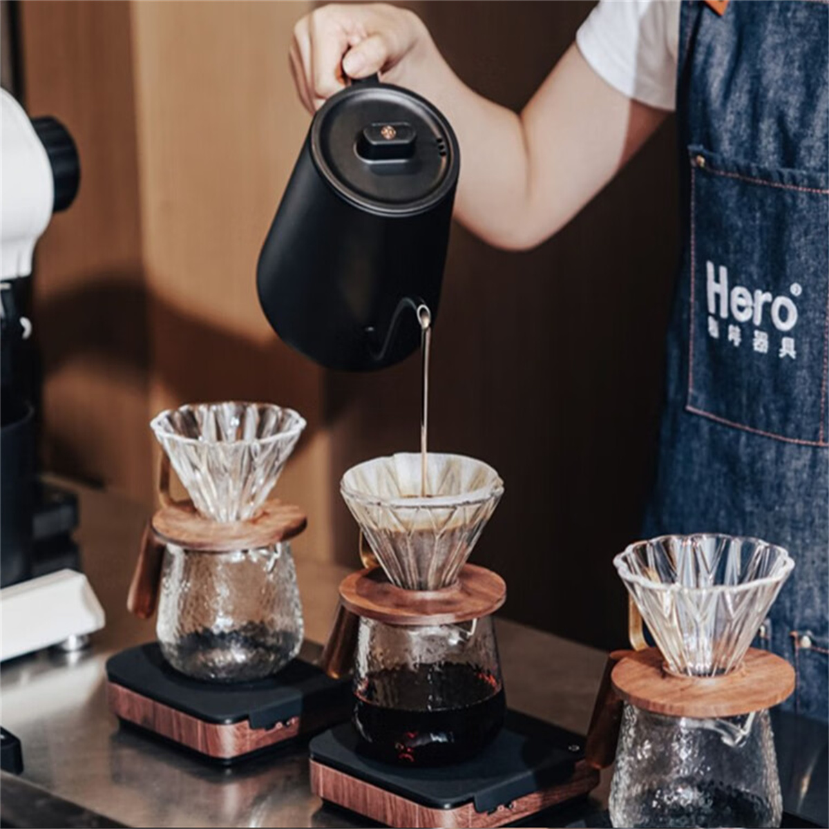 Eine Person gießt Kaffee aus einem schwarzen Wasserkocher in Glas-Pour-Over-Geräte. Die Szene ist ein Café.