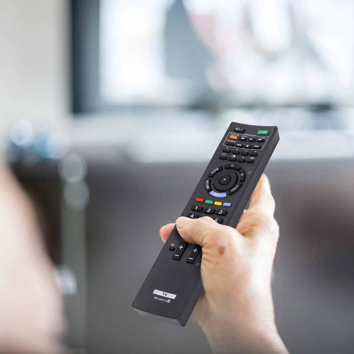 Eine Hand hält eine schwarze TV-Fernbedienung, die auf einen verschwommenen TV-Bildschirm im Hintergrund zeigt.