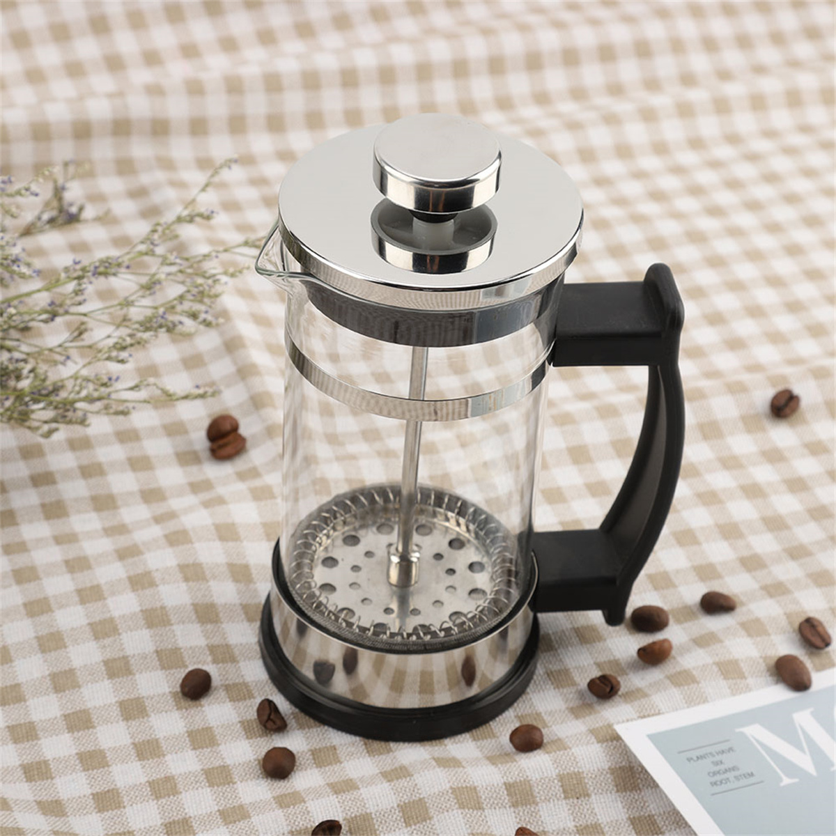 Glas-French-Press mit silbernem Deckel und schwarzem Griff. Kaffeebohnen drumherum.