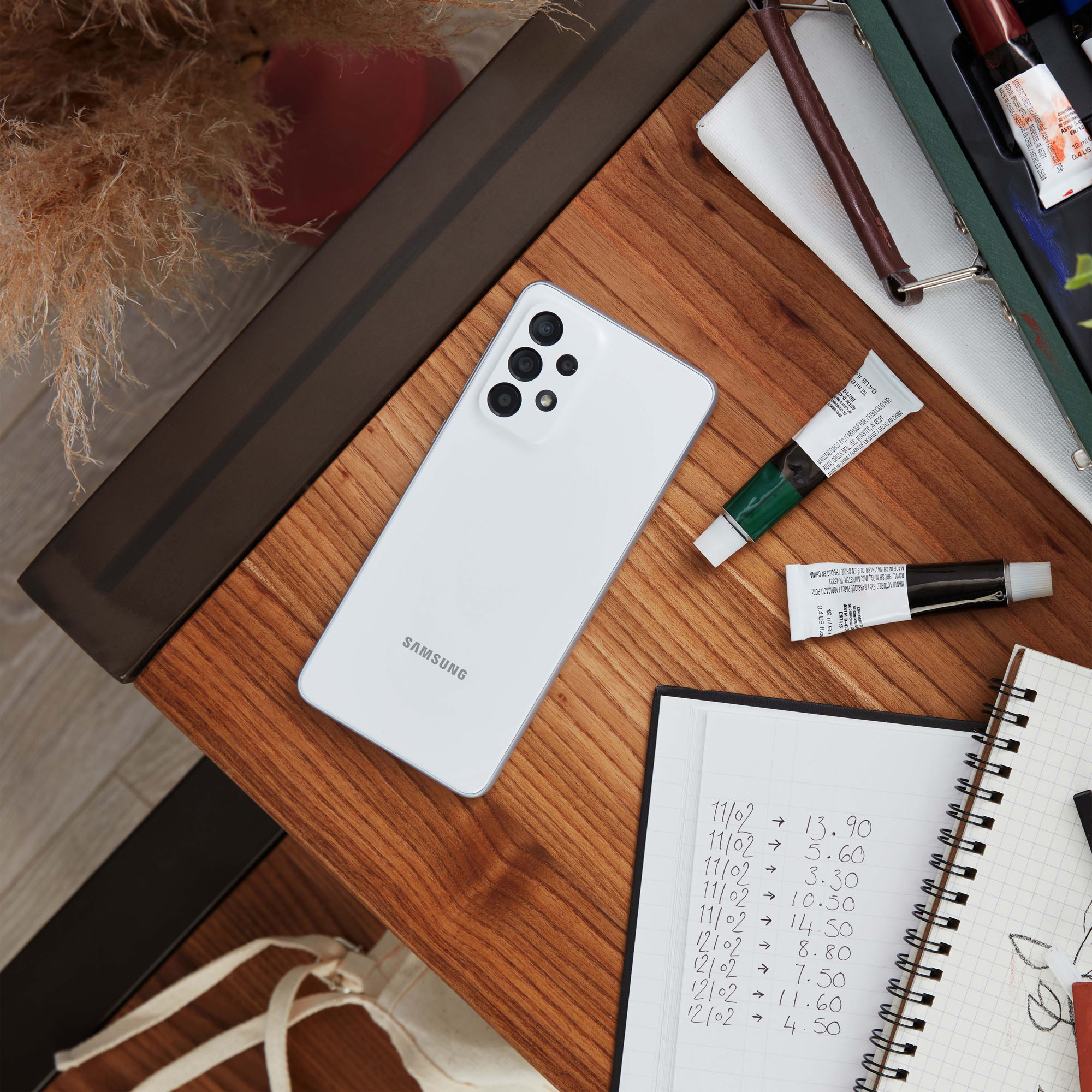 Biały telefon na drewnianym stole z tubkami farb, notatnikiem i innymi przedmiotami.