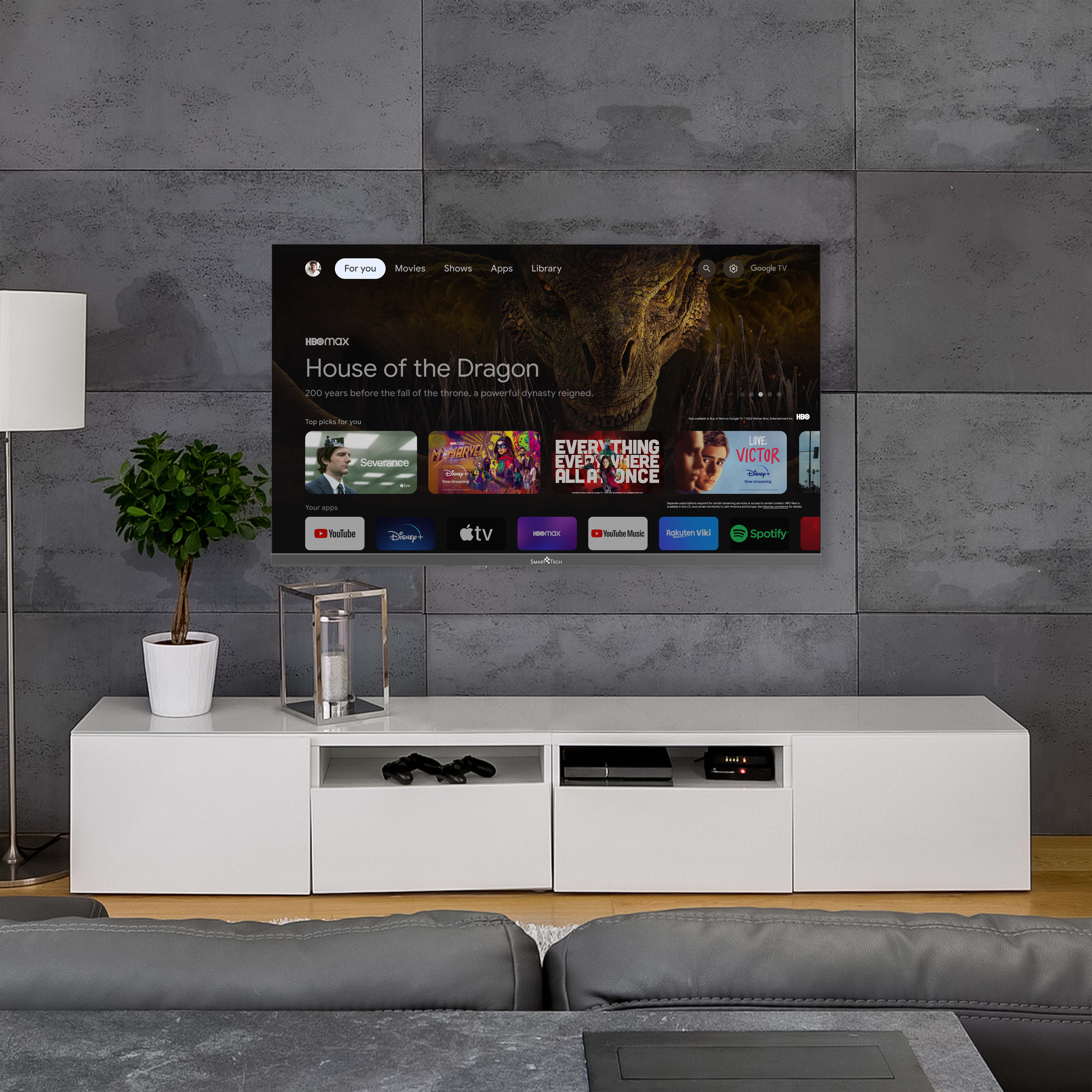 Telewizor wyświetla program. Biała konsola z rośliną i lampą. Szara sofa z przodu.