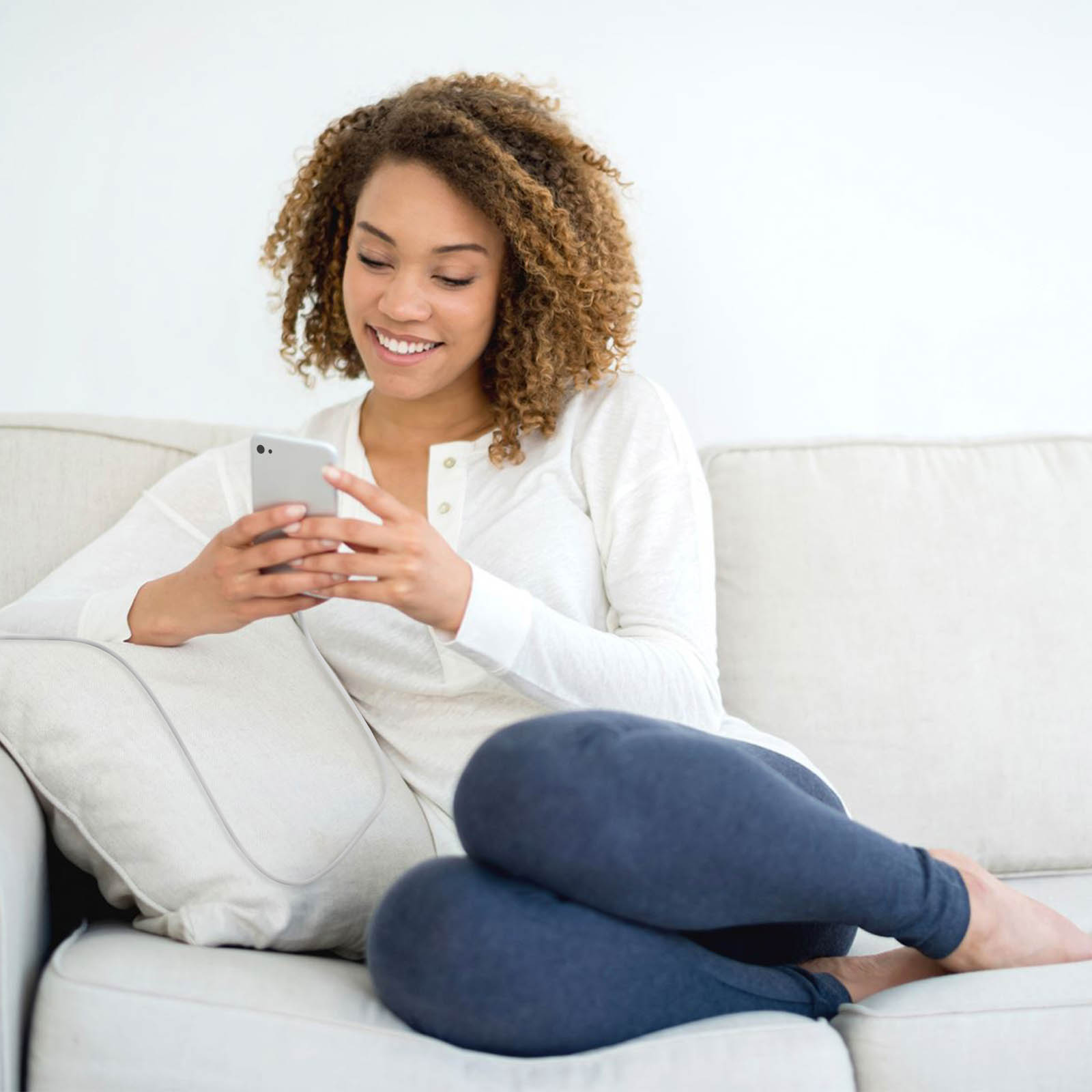 Eine lächelnde Frau mit lockigem Haar sitzt auf einem Sofa und schaut auf ihr Smartphone.