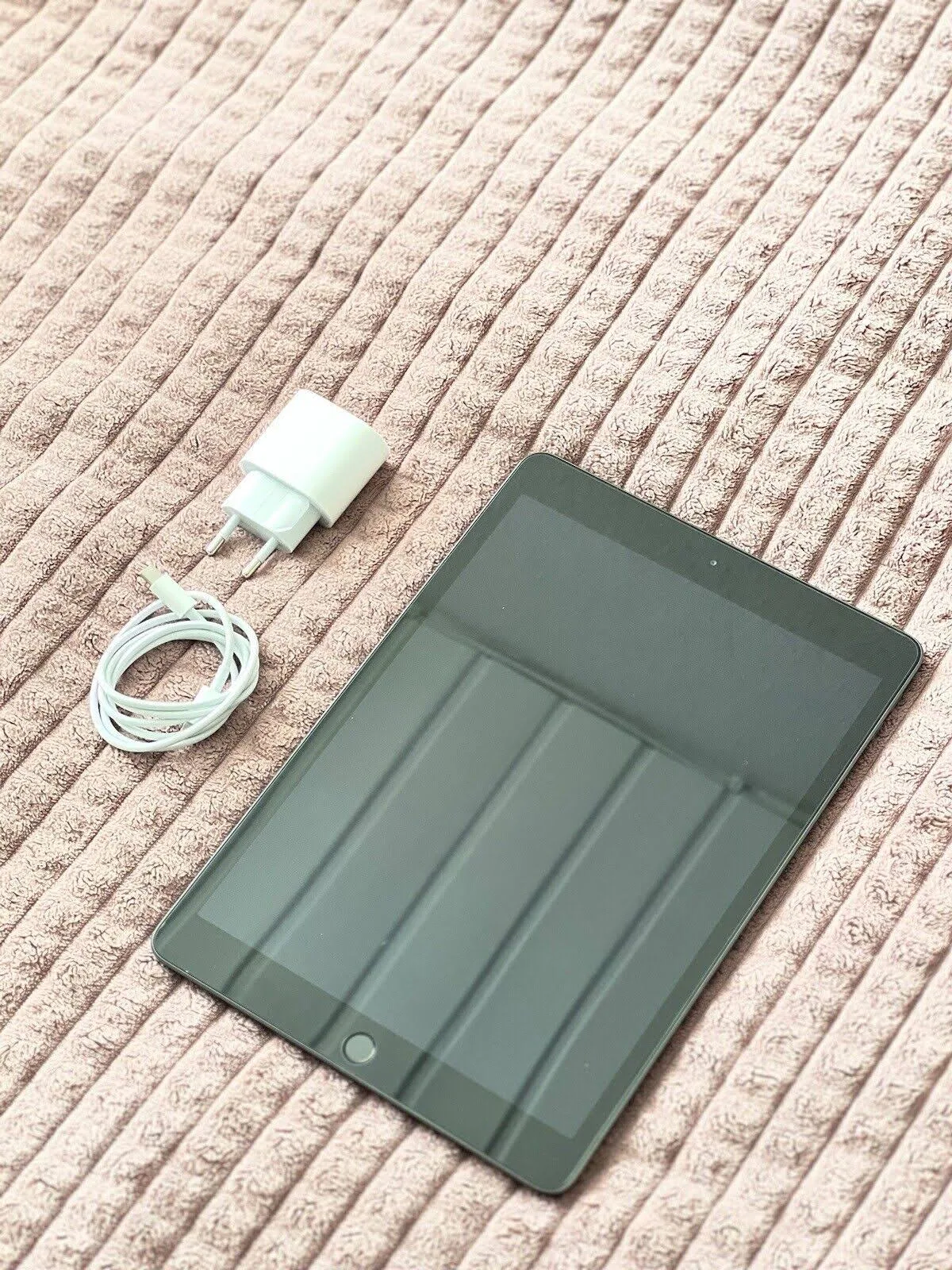 Tablet z ładowarką i kablem na różowej, teksturowanej powierzchni. Tablet jest czarny.