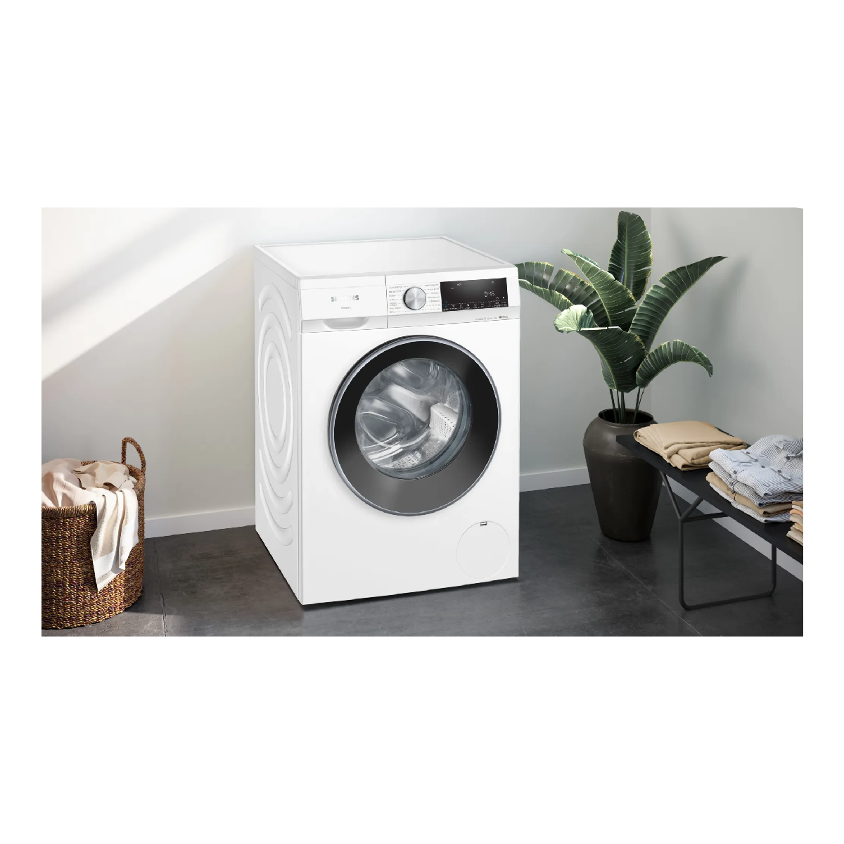 Una lavadora blanca con puerta negra y pantalla. Una cesta y una planta están cerca.
