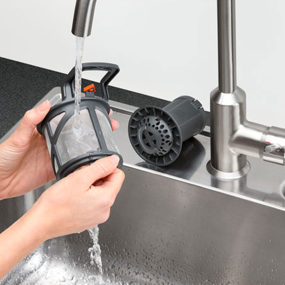 Mani che risciacquano un filtro per lavastoviglie sotto l'acqua corrente in un lavello da cucina.