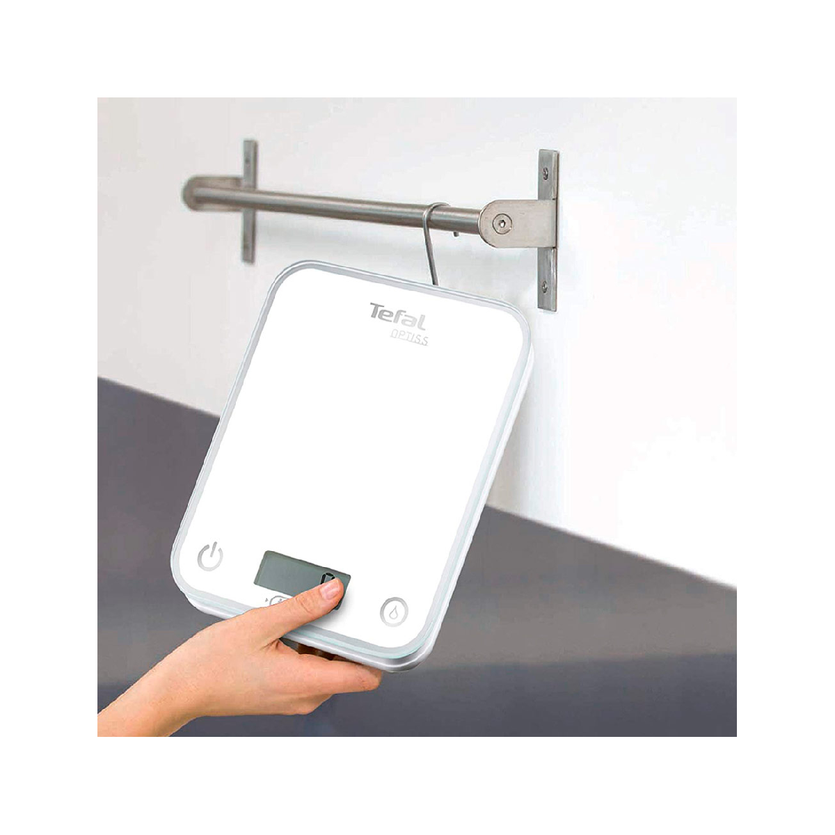 Dłoń trzyma biało-szarą wagę. Waga wisi na metalowym pręcie przymocowanym do ściany.