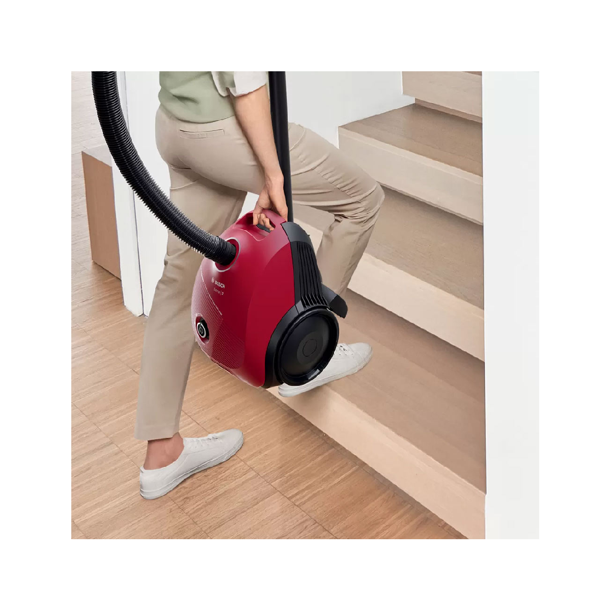 Osoba niosąca czerwony odkurzacz po drewnianych schodach. Beżowe spodnie, białe buty i czarny wąż.