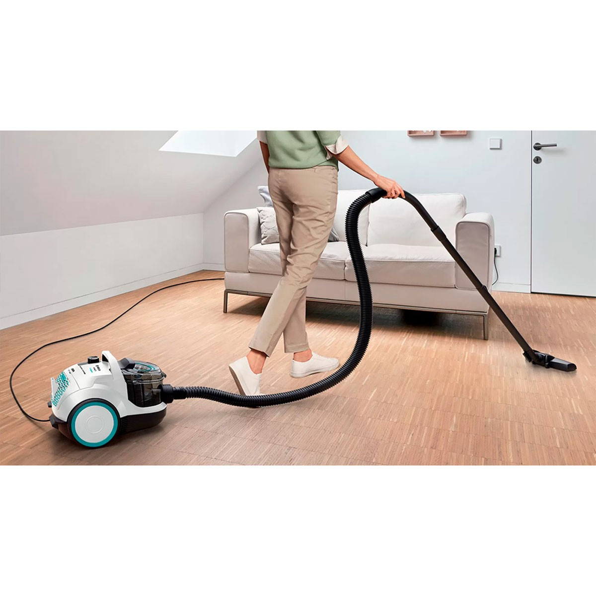 Osoba odkurza pokój. Biała sofa i drzwi w tle. Odkurzacz jest turkusowy i biały.