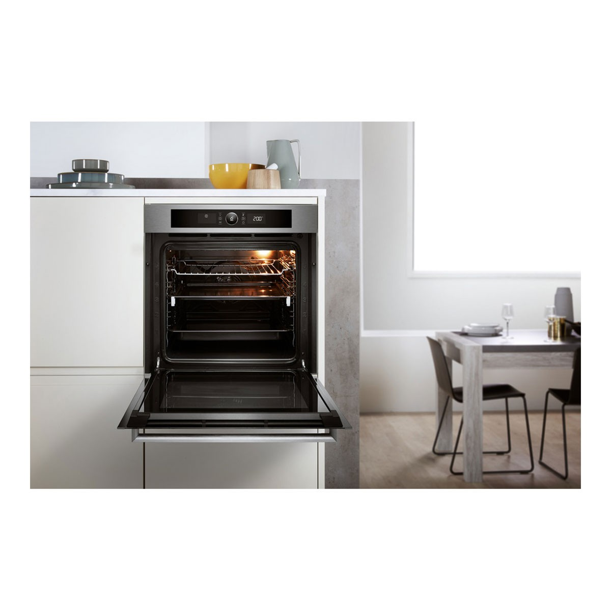 Horno con la puerta abierta. El interior muestra estantes. Zona de cocina con mesa y sillas. Fondo blanco.