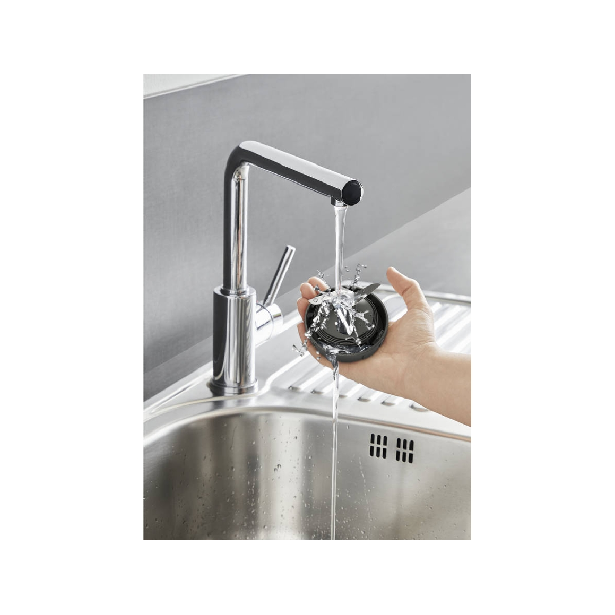 Una mano che tiene una lama di frullatore nera sotto l'acqua corrente in un lavello in acciaio inox.