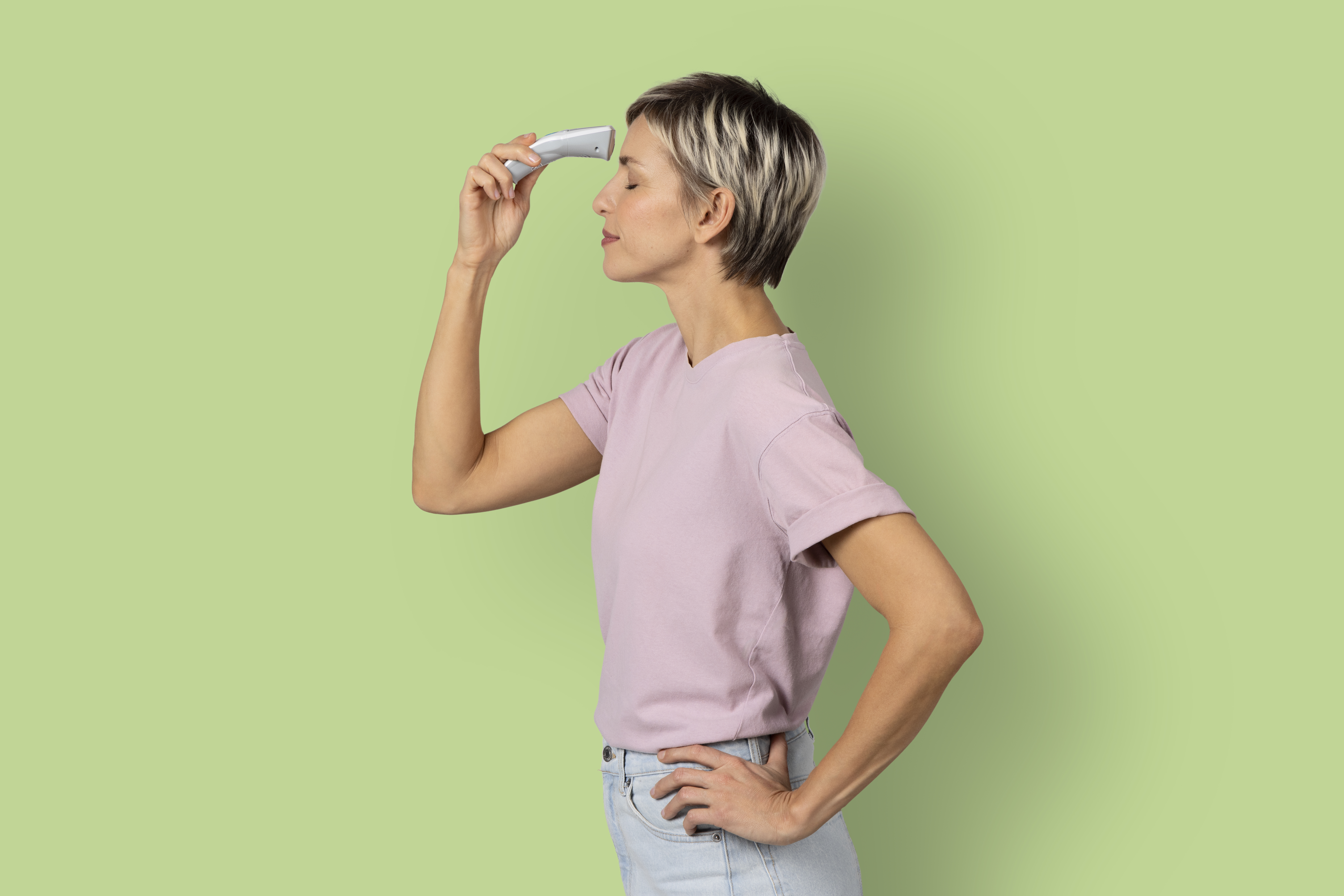 A woman using a digital thermometer on her forehead, standing against a green background.